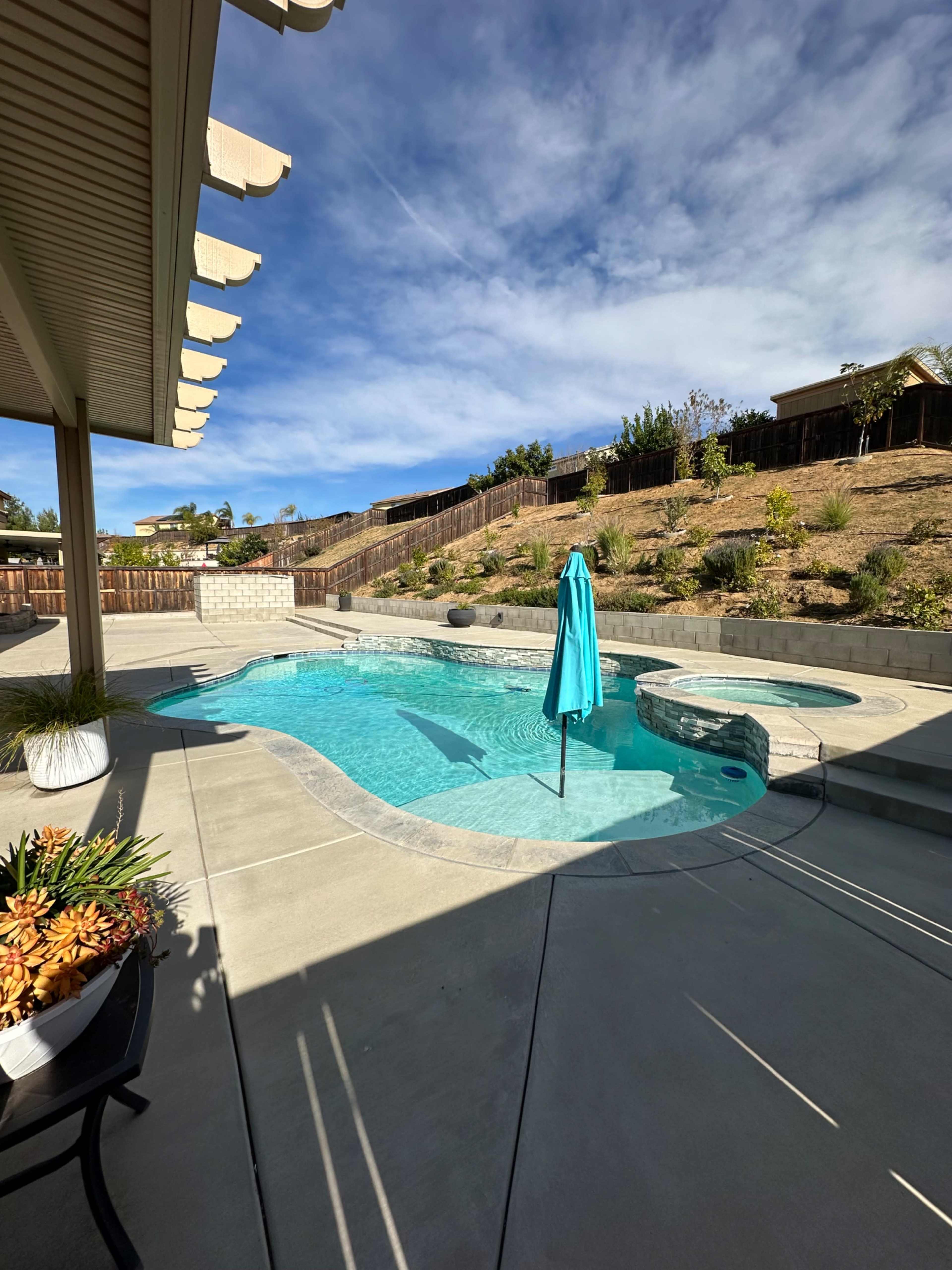 The image shows a backyard pool area featuring a shaped pool with a spa and a blue umbrella, surrounded by landscaping and a fenced area.