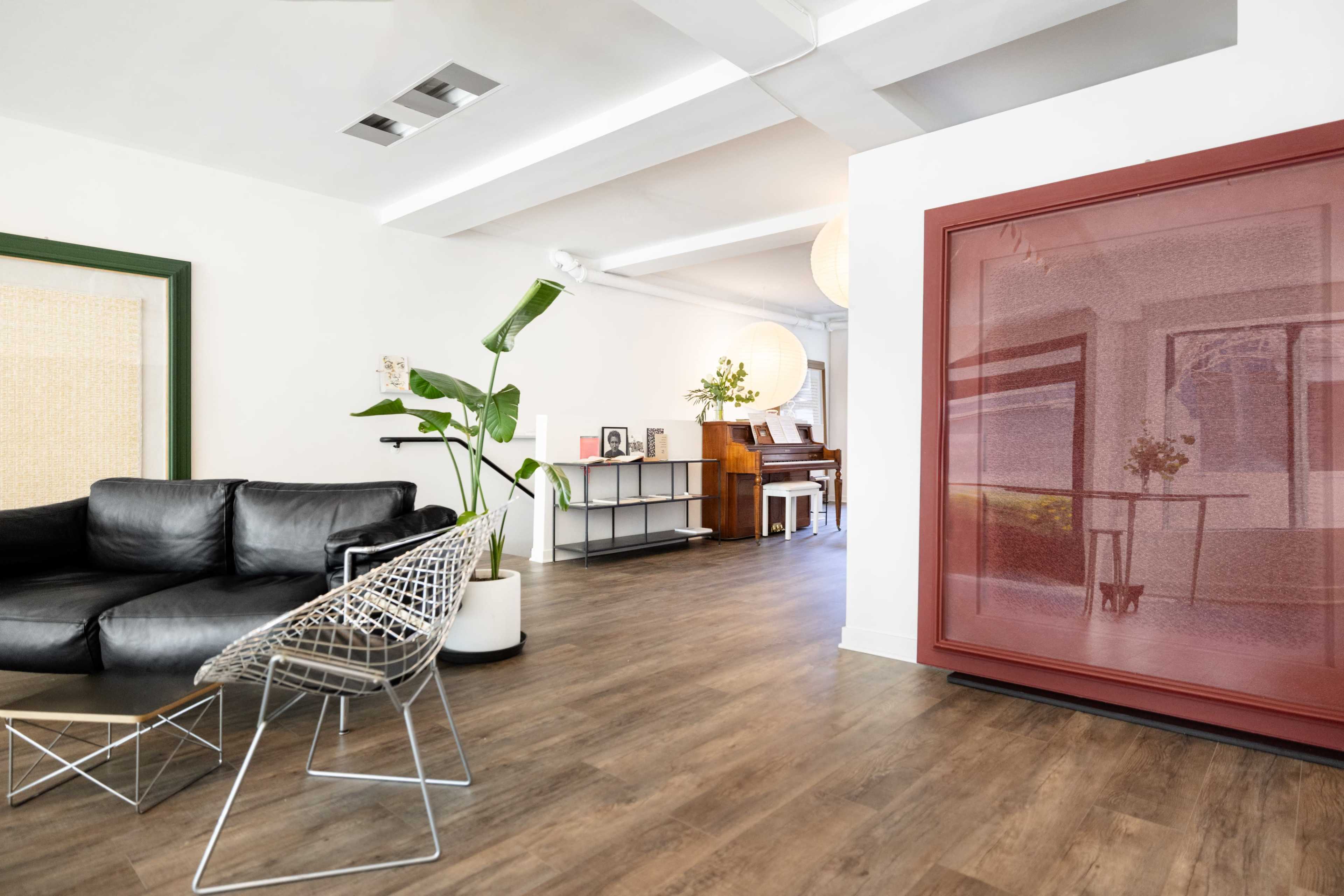 A modern living space featuring a black leather sofa, a wire-frame chair, a wooden floor, and a large red-framed artwork, with a glimpse of a piano in the background.