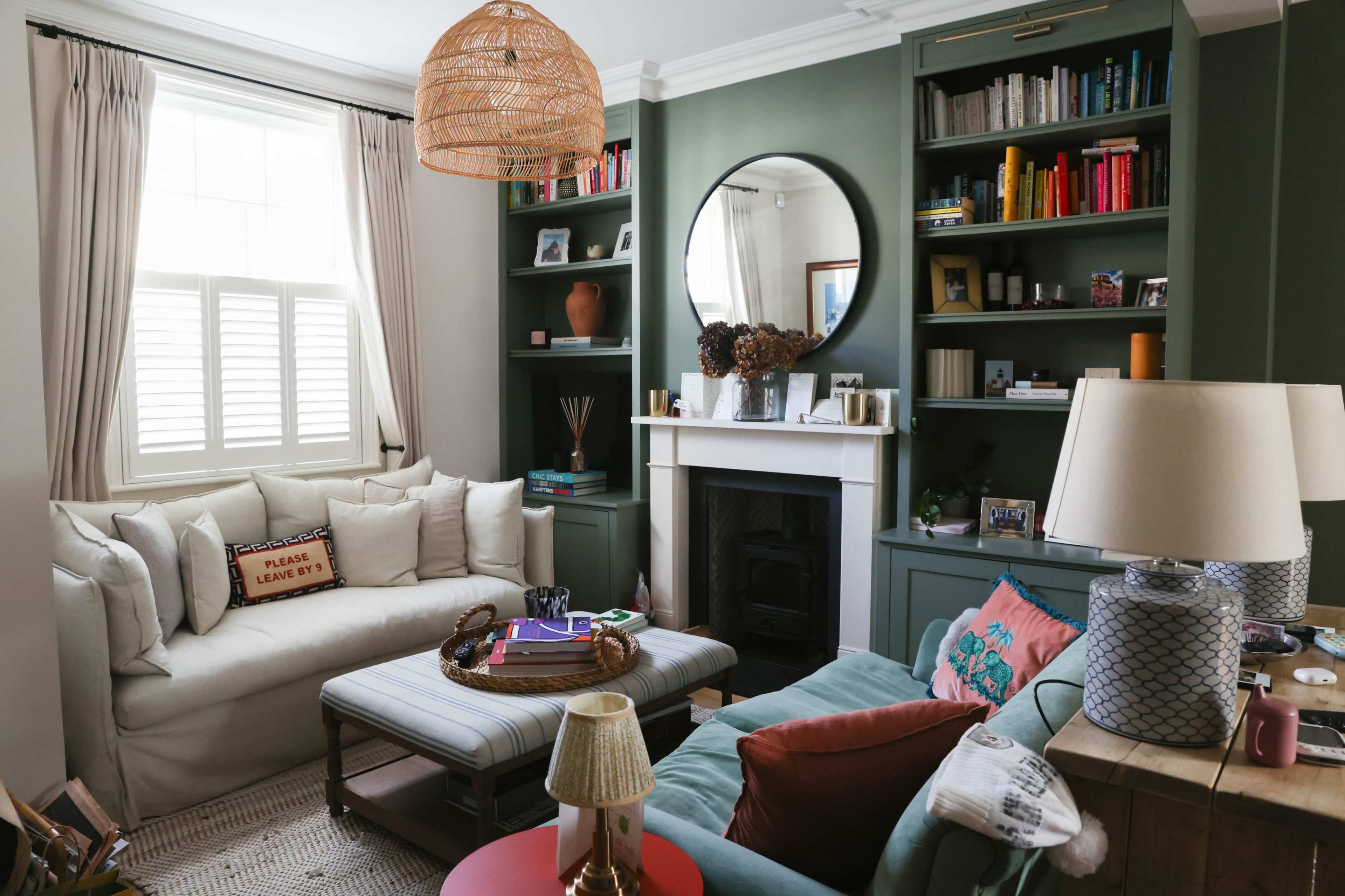 The image shows a living room with a light-colored sofa, a teal armchair, a round mirror above a fireplace, and shelves filled with books and decorative items.
