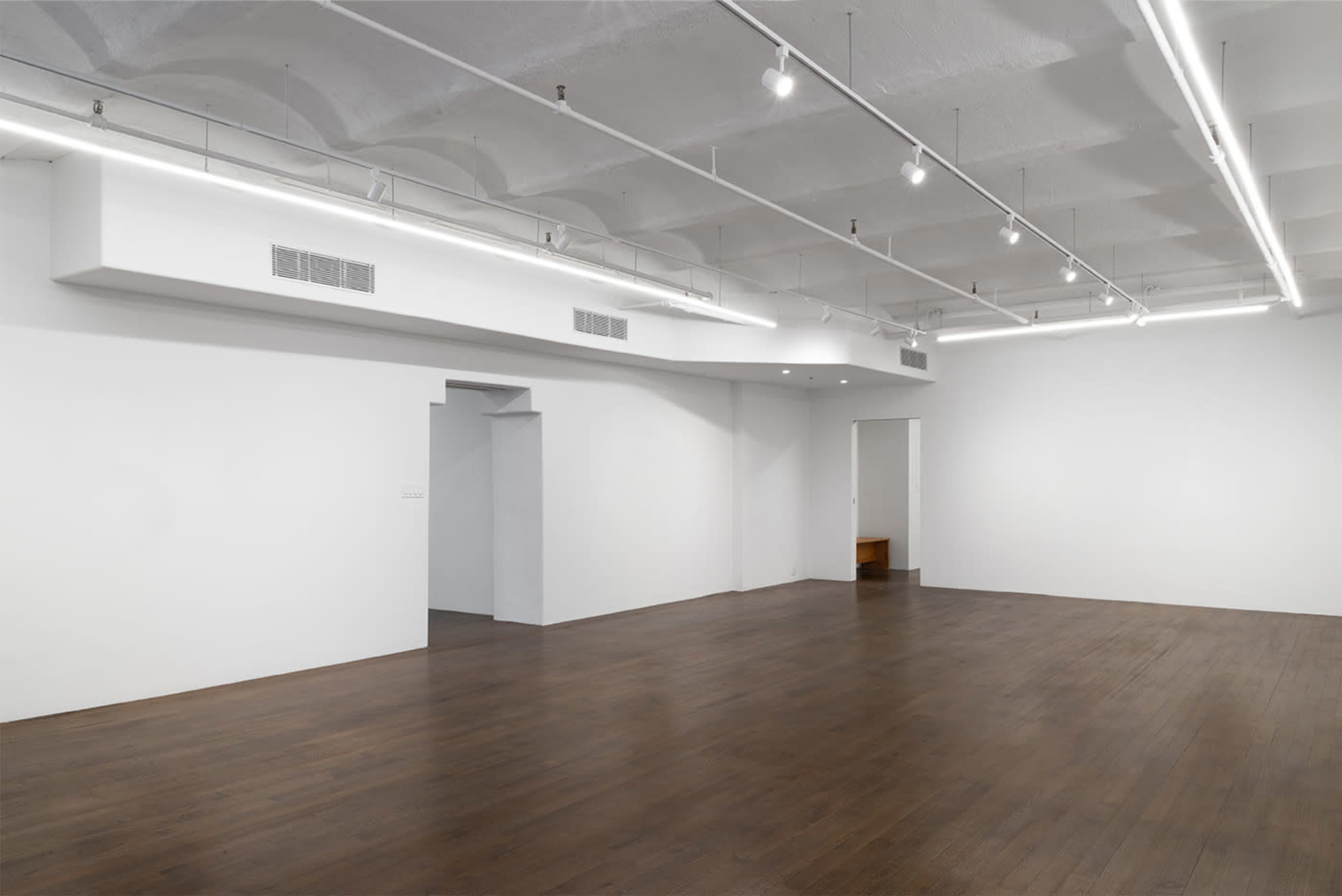 The image shows a spacious, empty room with wooden flooring and white walls, featuring overhead lighting and a doorway on the left side.