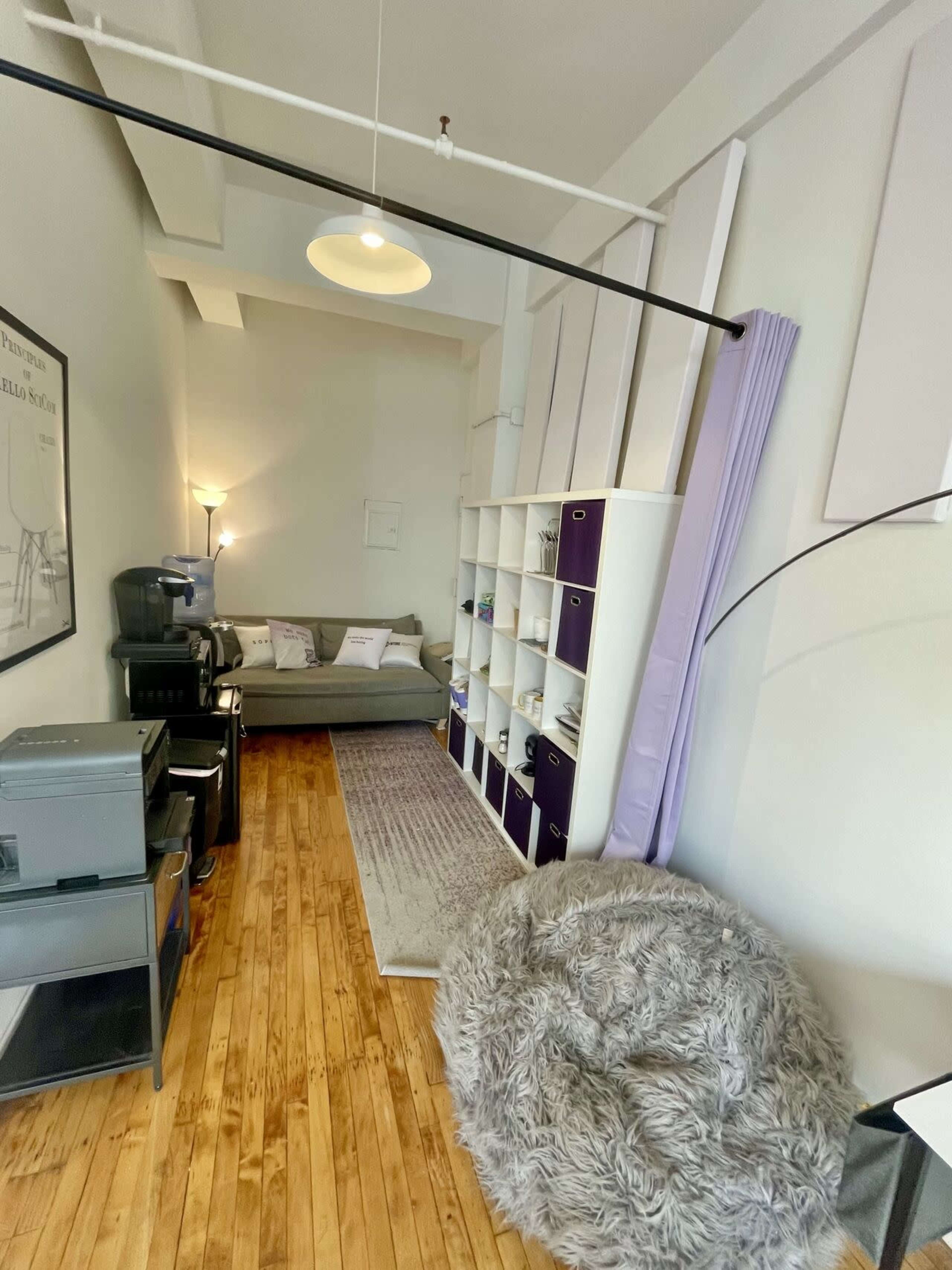 The image shows a narrow room featuring a gray sofa, a large storage unit, a printer, and a cozy bean bag chair on a wooden floor.