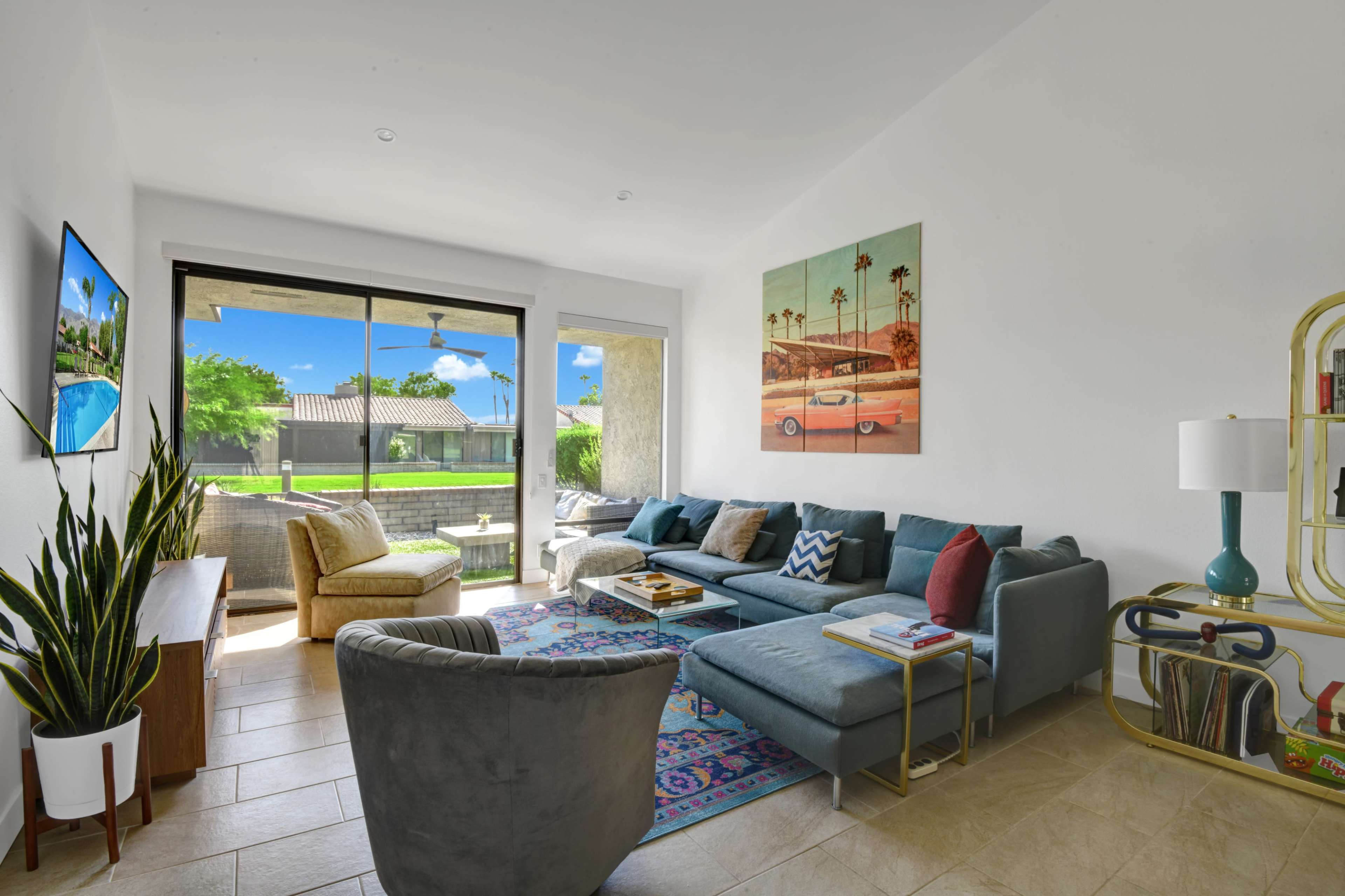 The scene shows a modern living room featuring a large sectional sofa, a patterned area rug, and a sliding glass door that opens to a patio with a view of green grass and palm trees.