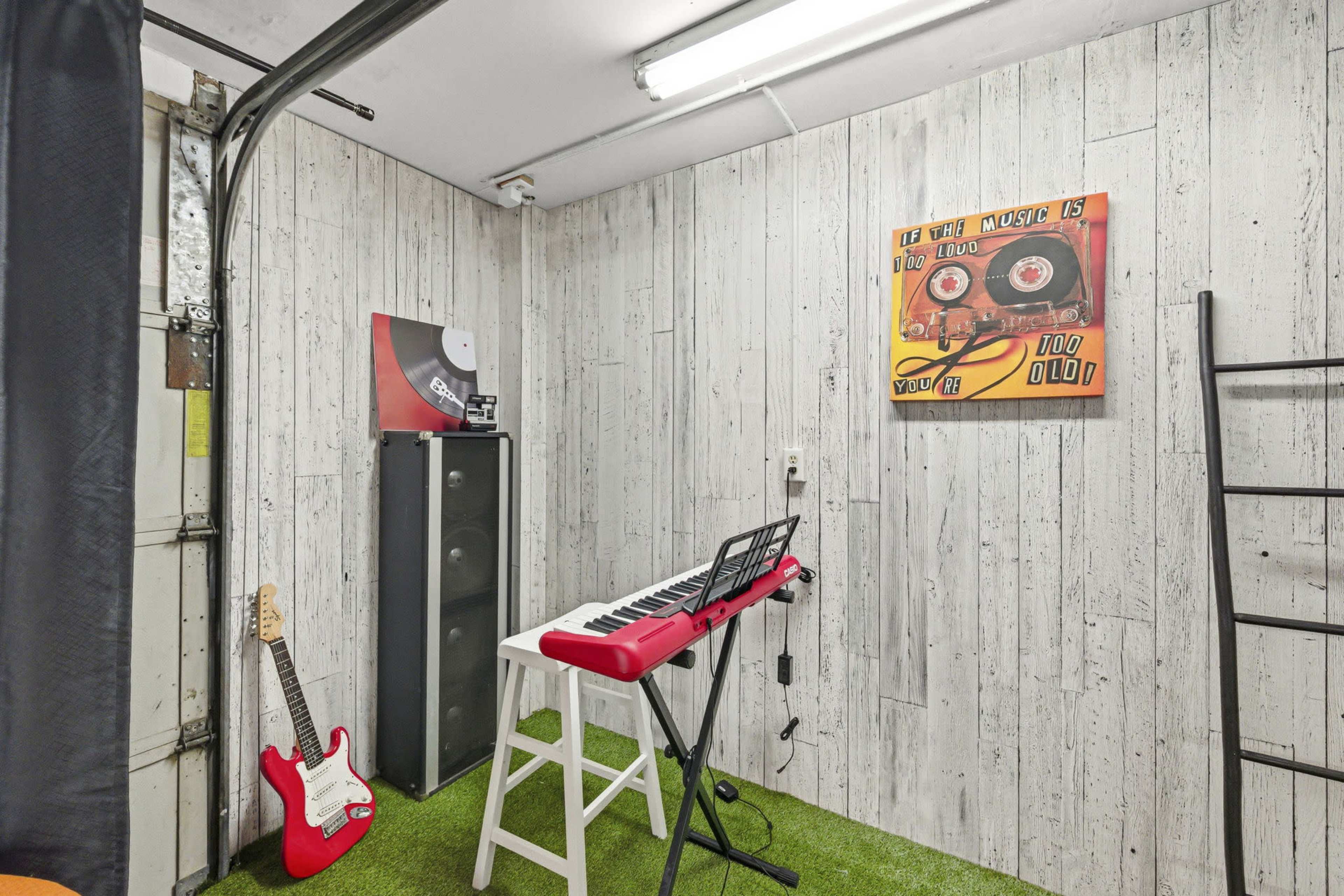 The image shows a small music practice space with a keyboard on a stool, a guitar leaning against the wall, and sound equipment next to it.