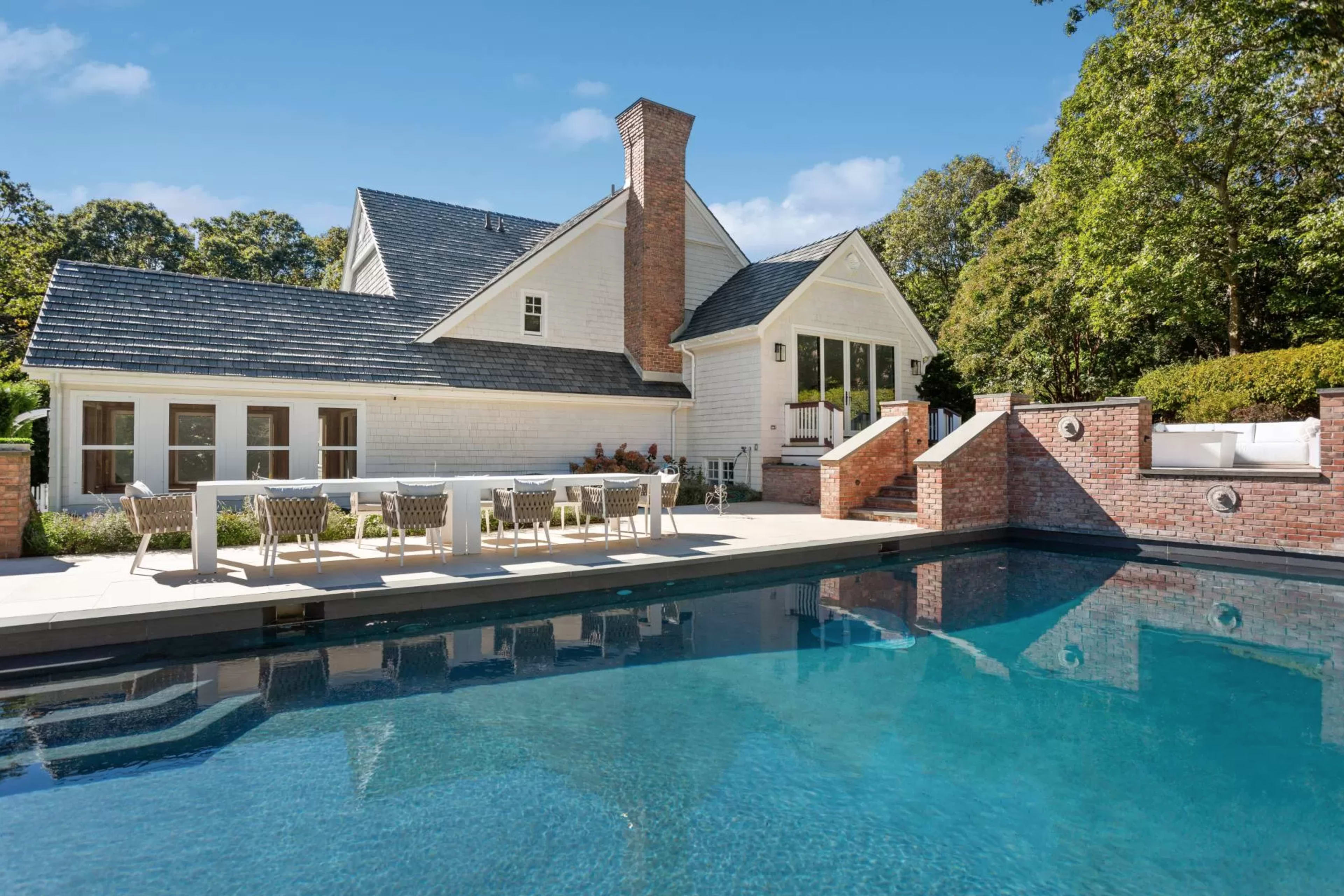 A modern house with a large swimming pool and an outdoor dining area is set against a backdrop of trees.