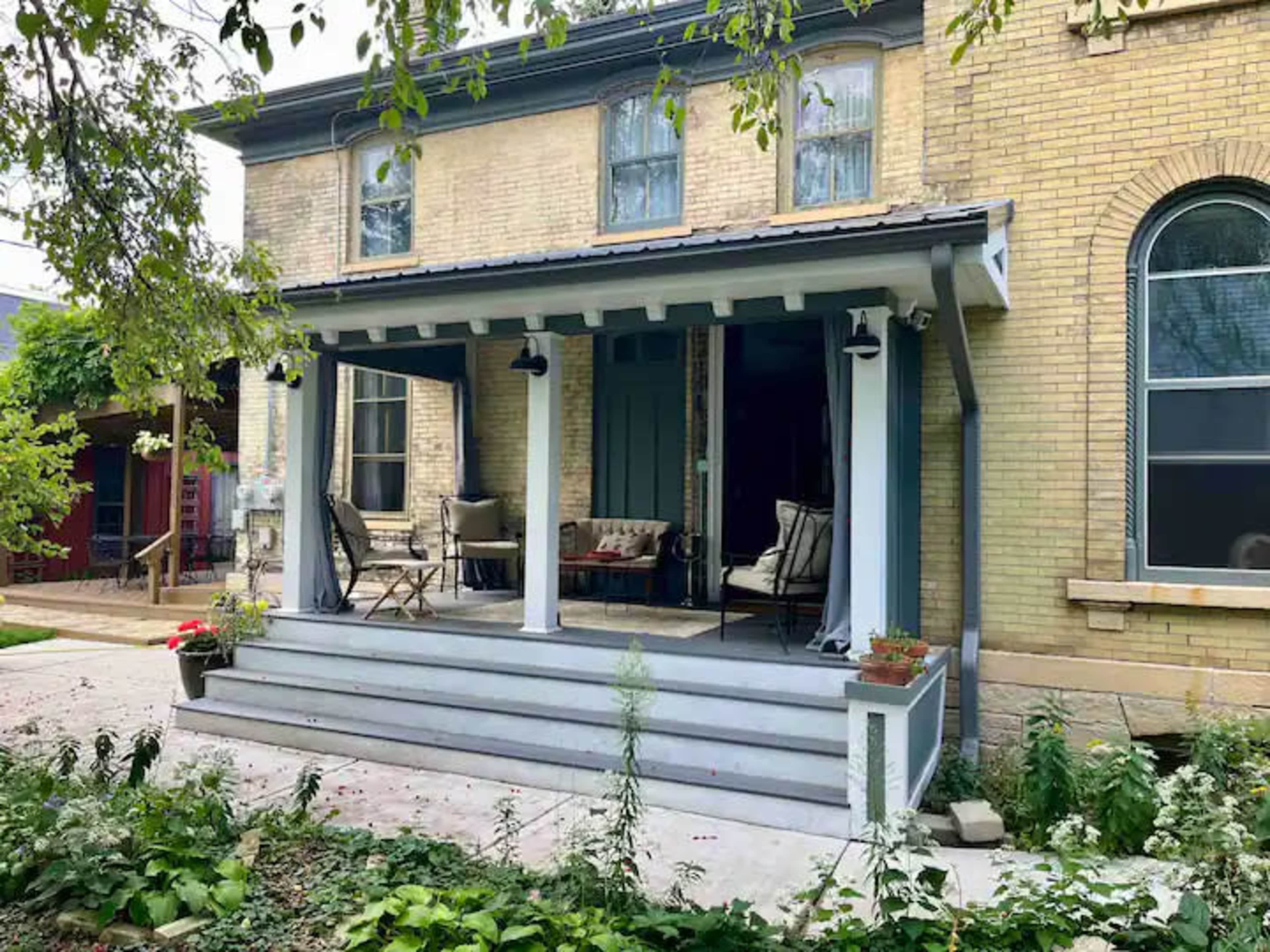 A two-story house with a porch featuring seating and potted plants, surrounded by greenery and a paved pathway.