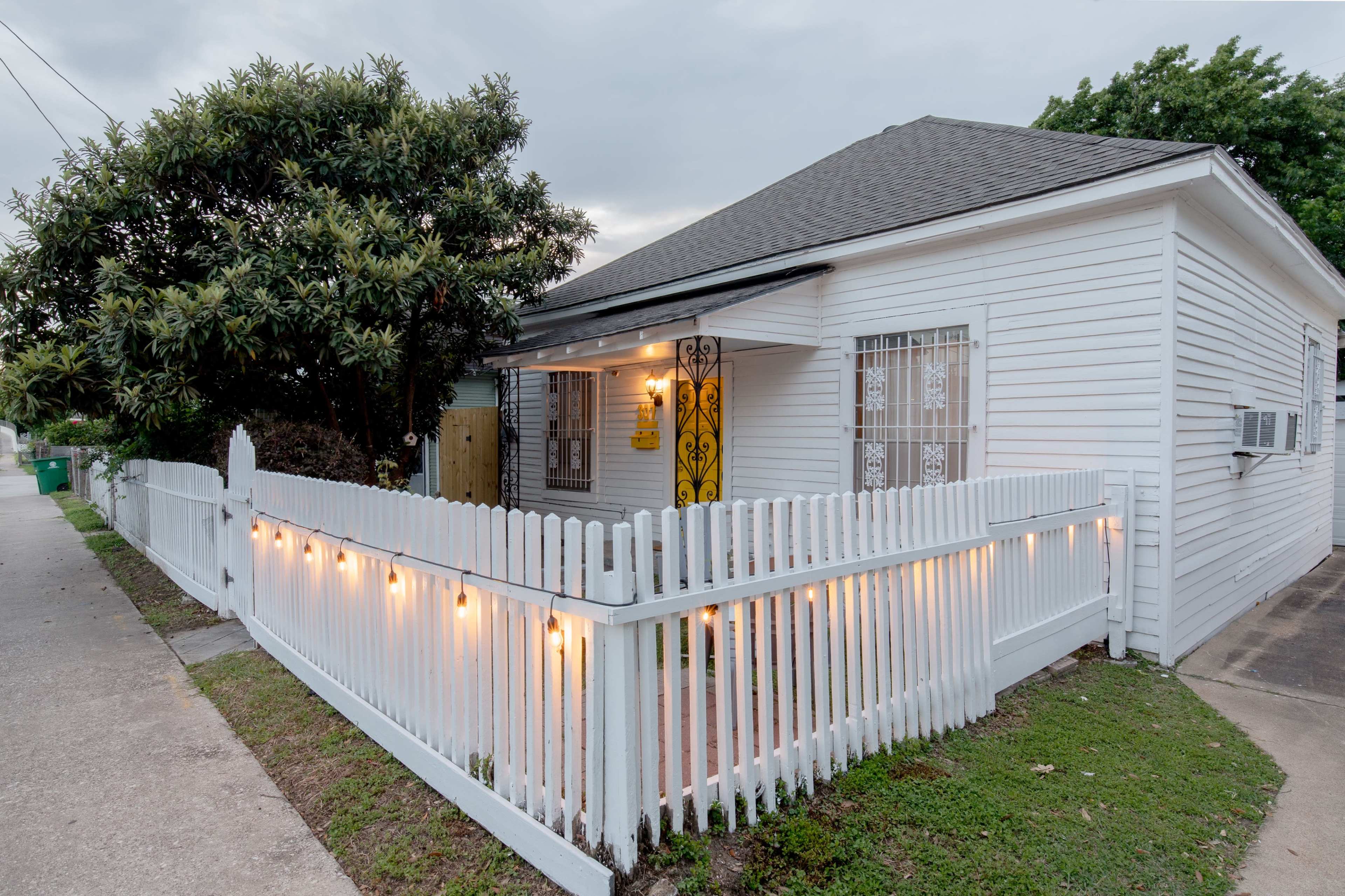 A white wooden house with a decorative front door is enclosed by a white picket fence, illuminated by string lights, and is surrounded by greenery.
