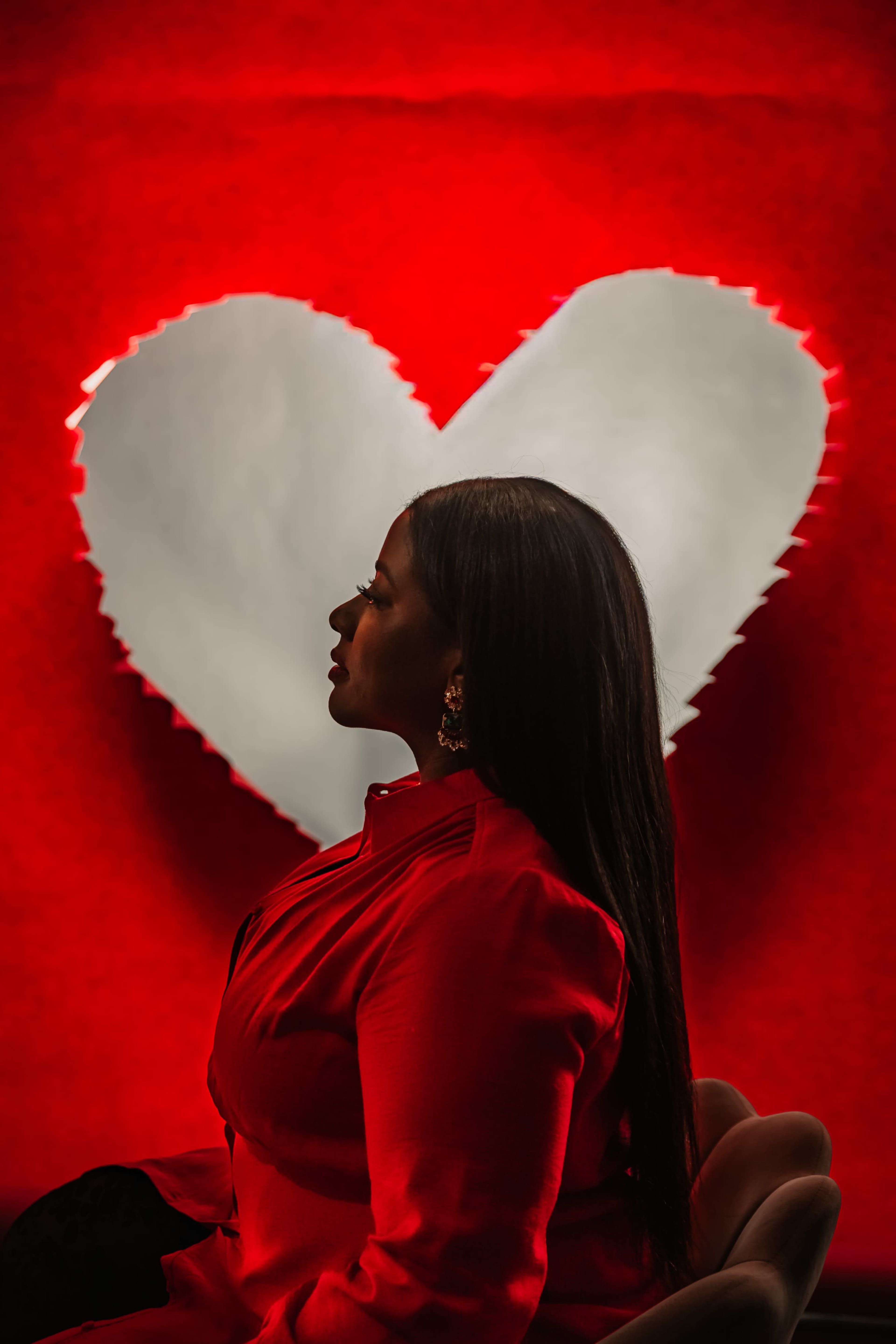 A woman in a red blouse sits in profile in front of a large heart-shaped background.