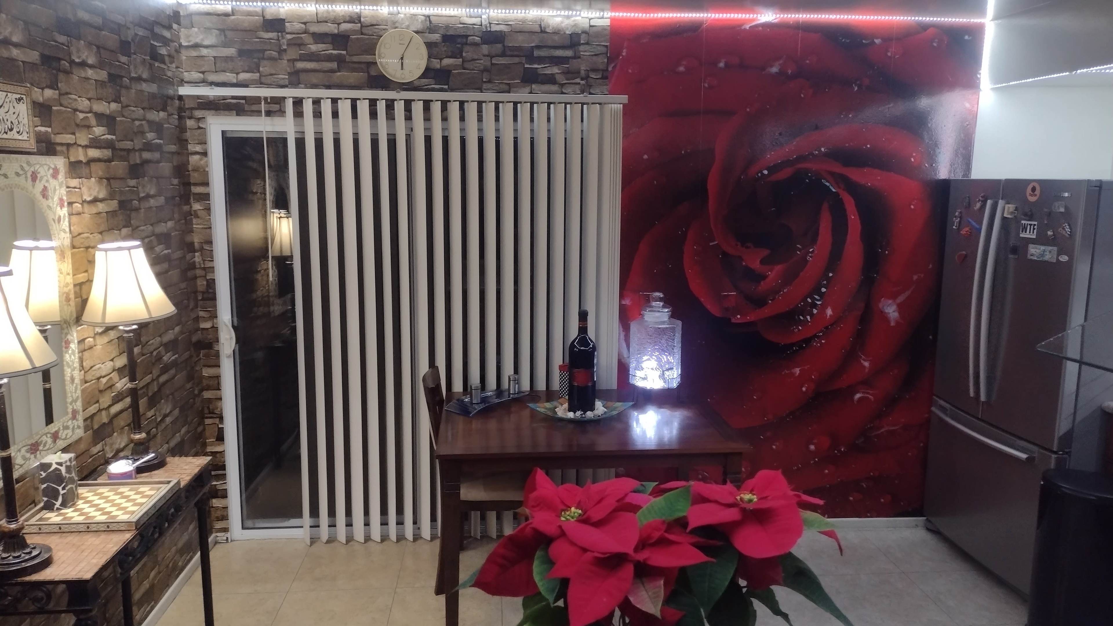 A dining area with a table set for drinks, a decorative wall featuring a large red rose, and a window with vertical blinds.