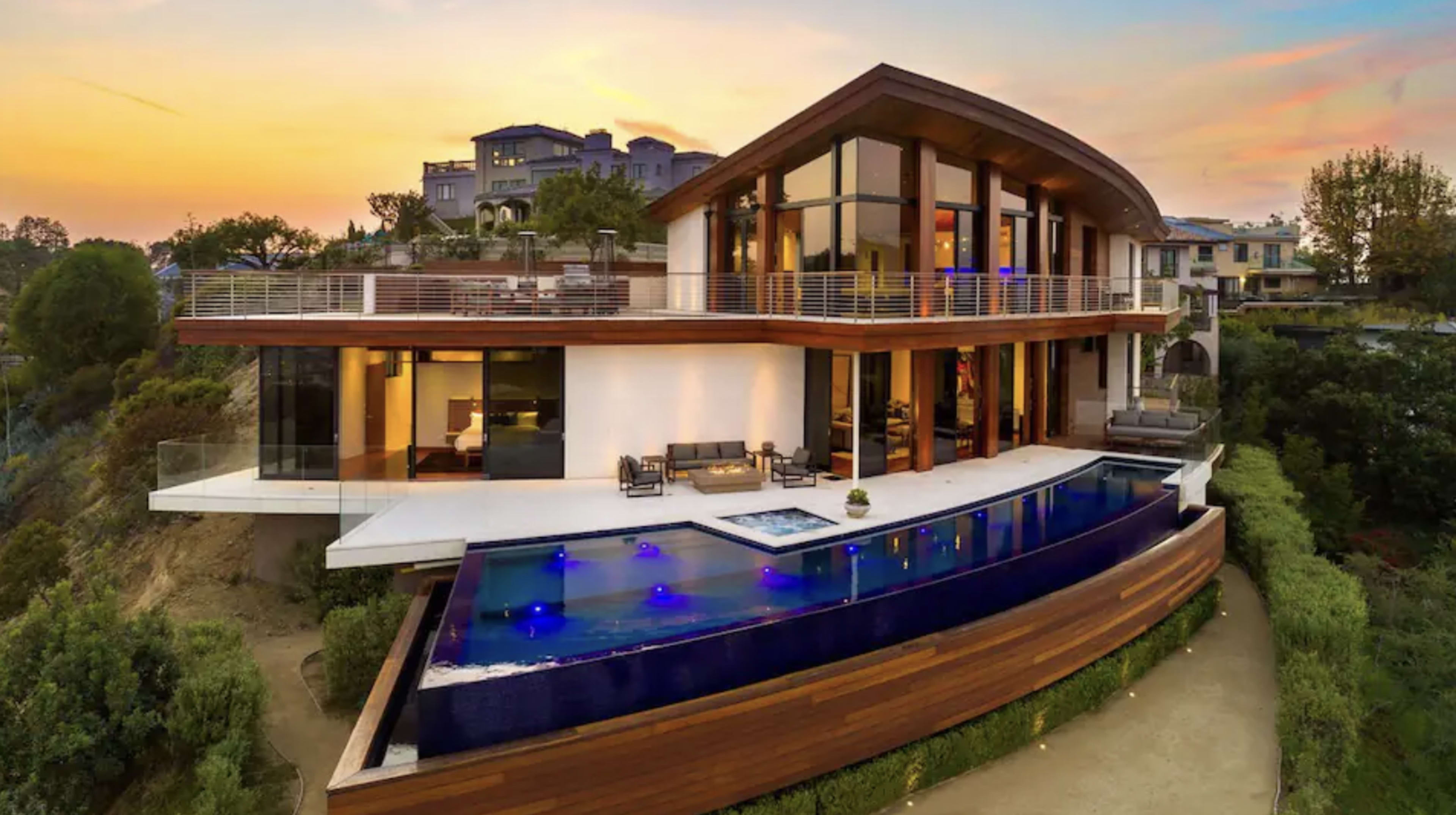 A modern house with large windows, featuring a sleek swimming pool that extends toward the edge of a hillside, all set against a sunset sky.