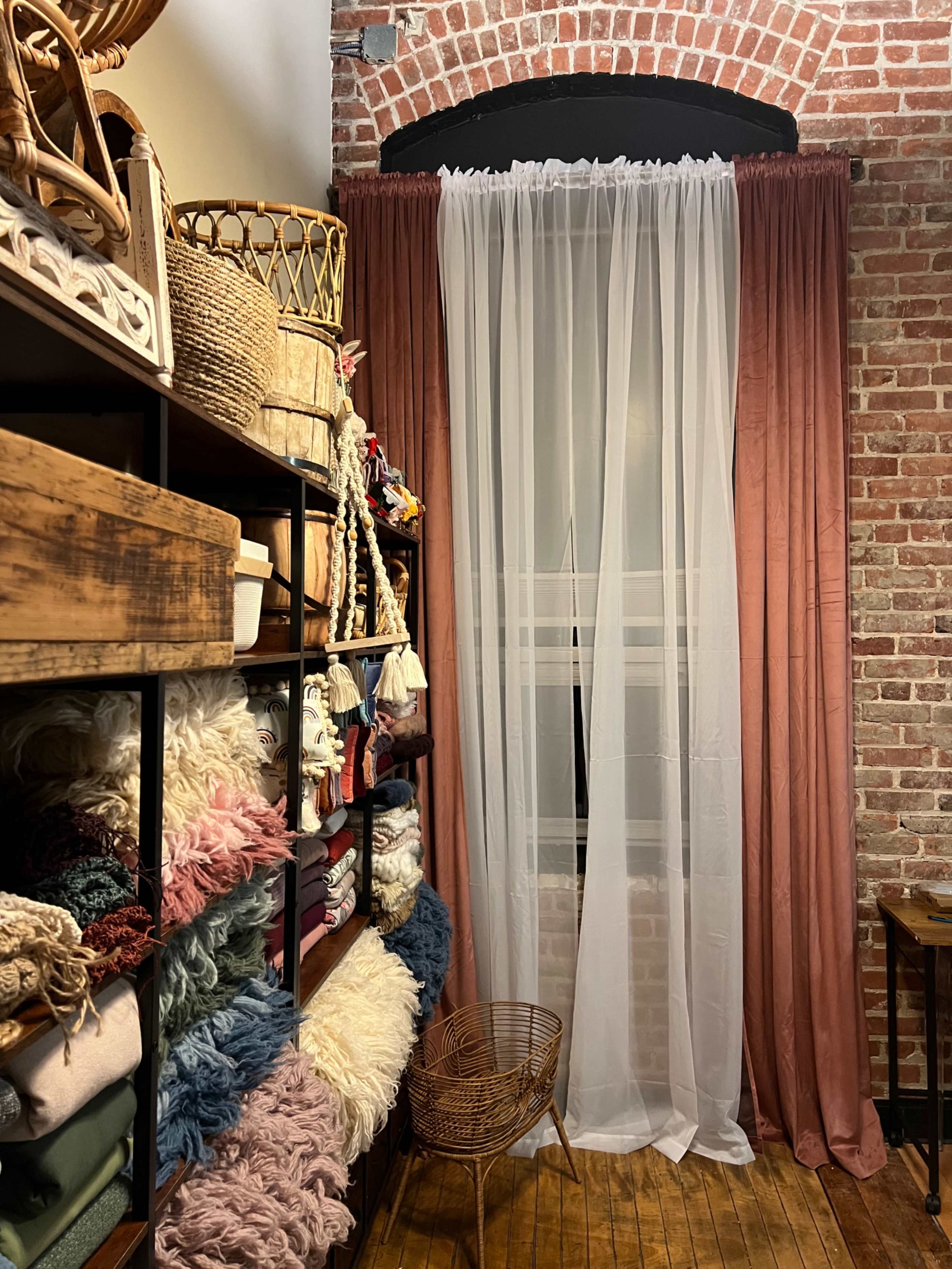 The image shows a cozy room with a window dressed in sheer white curtains, surrounded by exposed brick walls and shelves filled with colorful textiles and woven baskets.