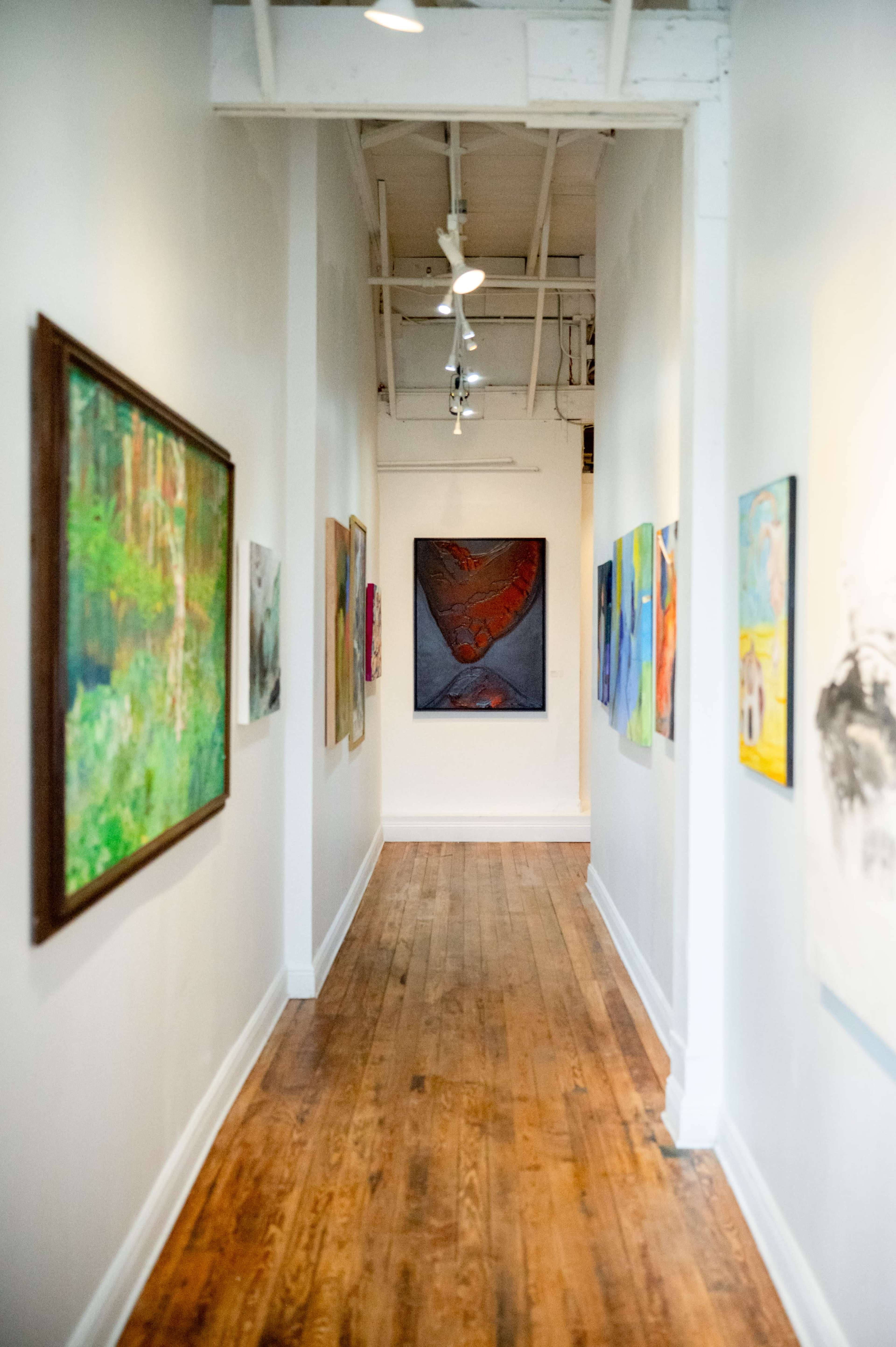 The image shows a long, narrow gallery hallway lined with various colorful artworks on the walls.
