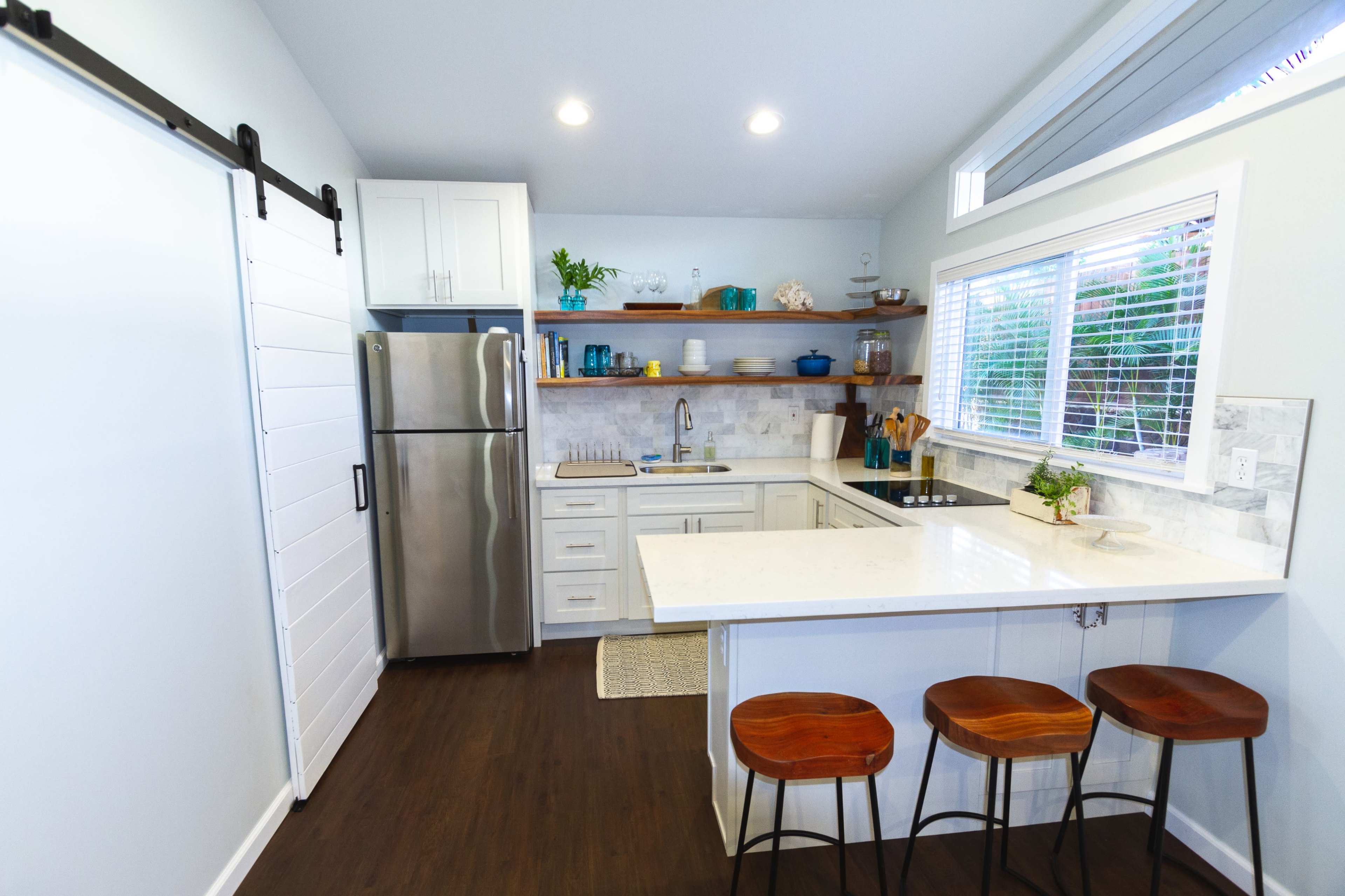 A compact kitchen features stainless steel appliances, white cabinetry, and a small dining area with three wooden stools.