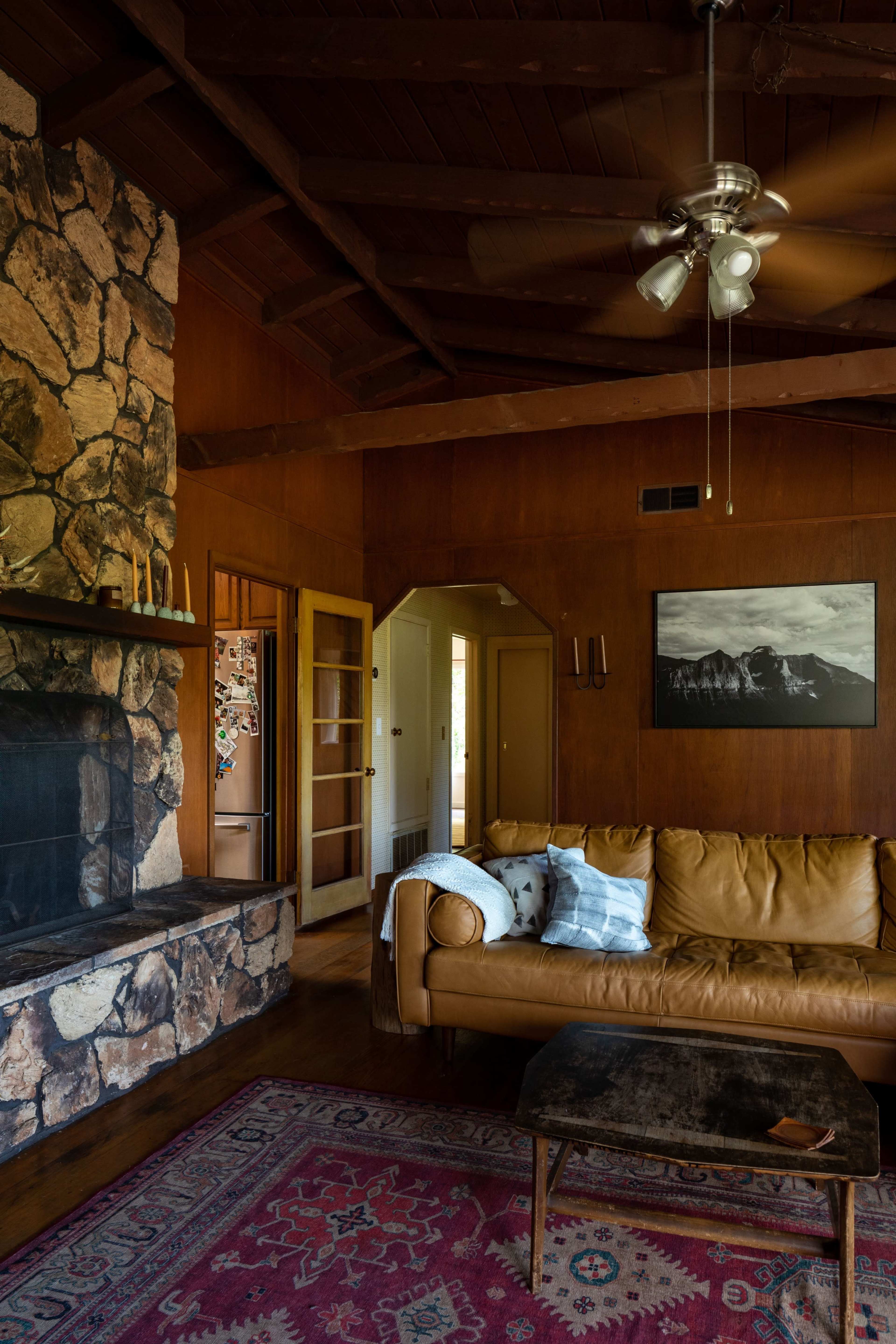A living room with a stone fireplace, a brown leather sofa, a wooden coffee table, and a mountain landscape photograph on the wall, all under a ceiling fan.