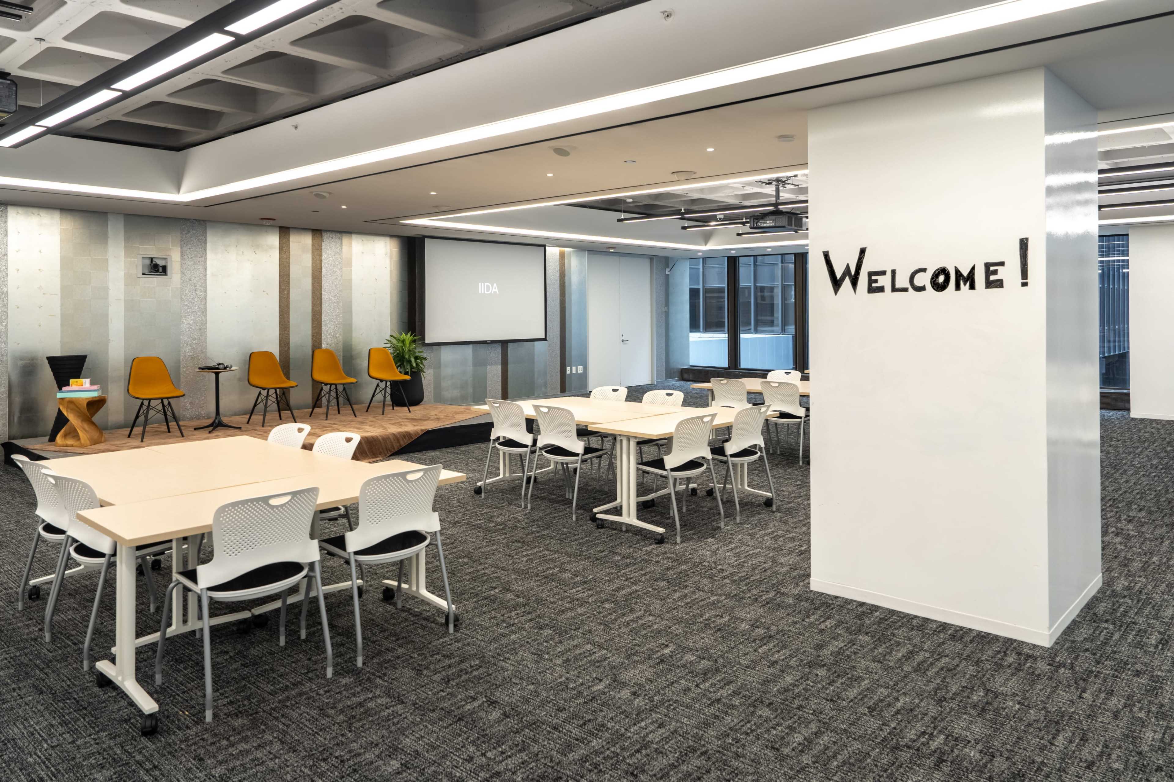 The image shows a modern office space with tables and chairs arranged for meetings, a presentation screen, and a welcoming sign on the wall.