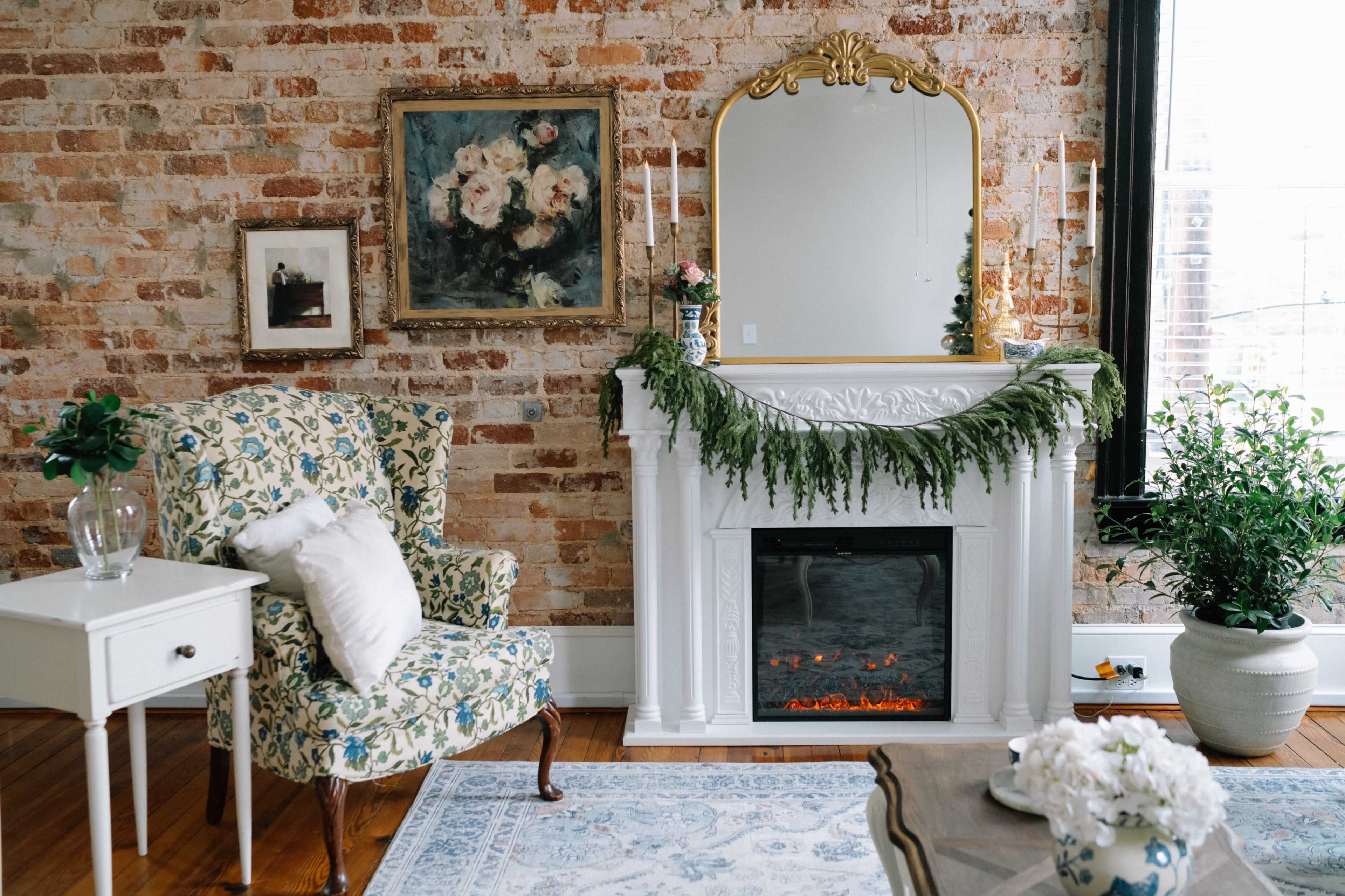 The room features a brick wall with a white mantel decorated with greenery, a mirror above it, a patterned armchair, a side table, and a cozy area rug.