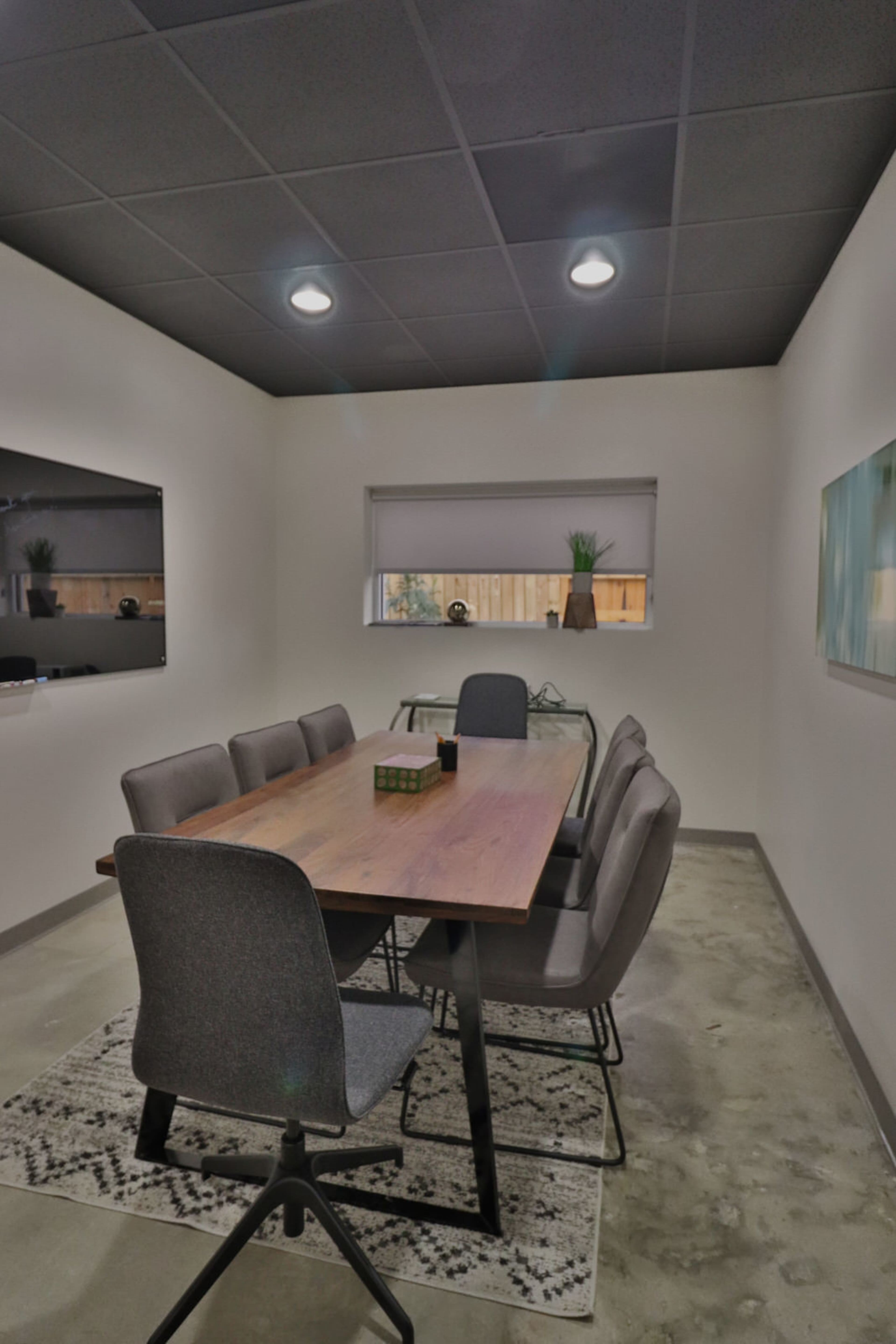 A modern conference room features a large wooden table surrounded by gray chairs, with a television mounted on the wall and large windows allowing natural light.