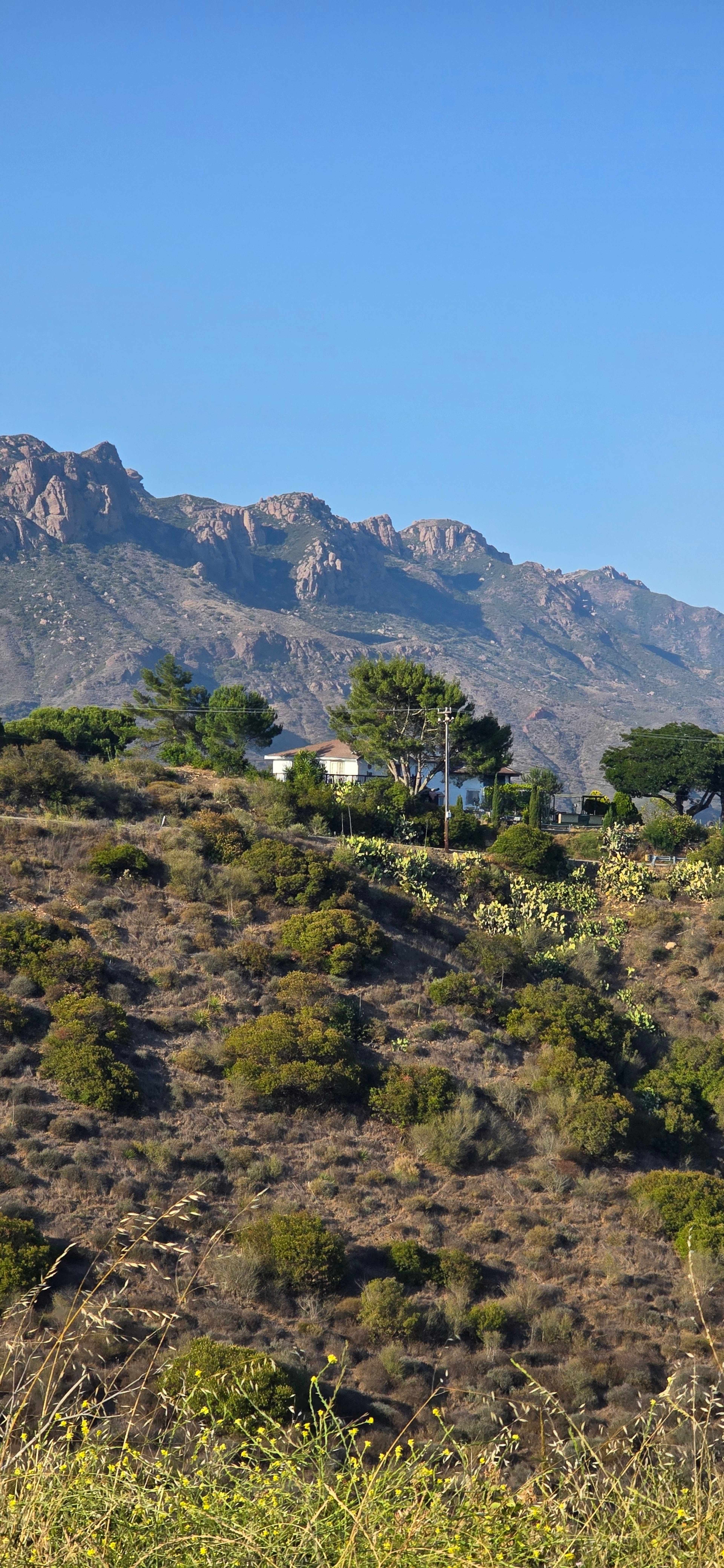 The image shows a hillside with sparse vegetation and a white house surrounded by trees, set against a backdrop of rocky mountains under a clear blue sky.