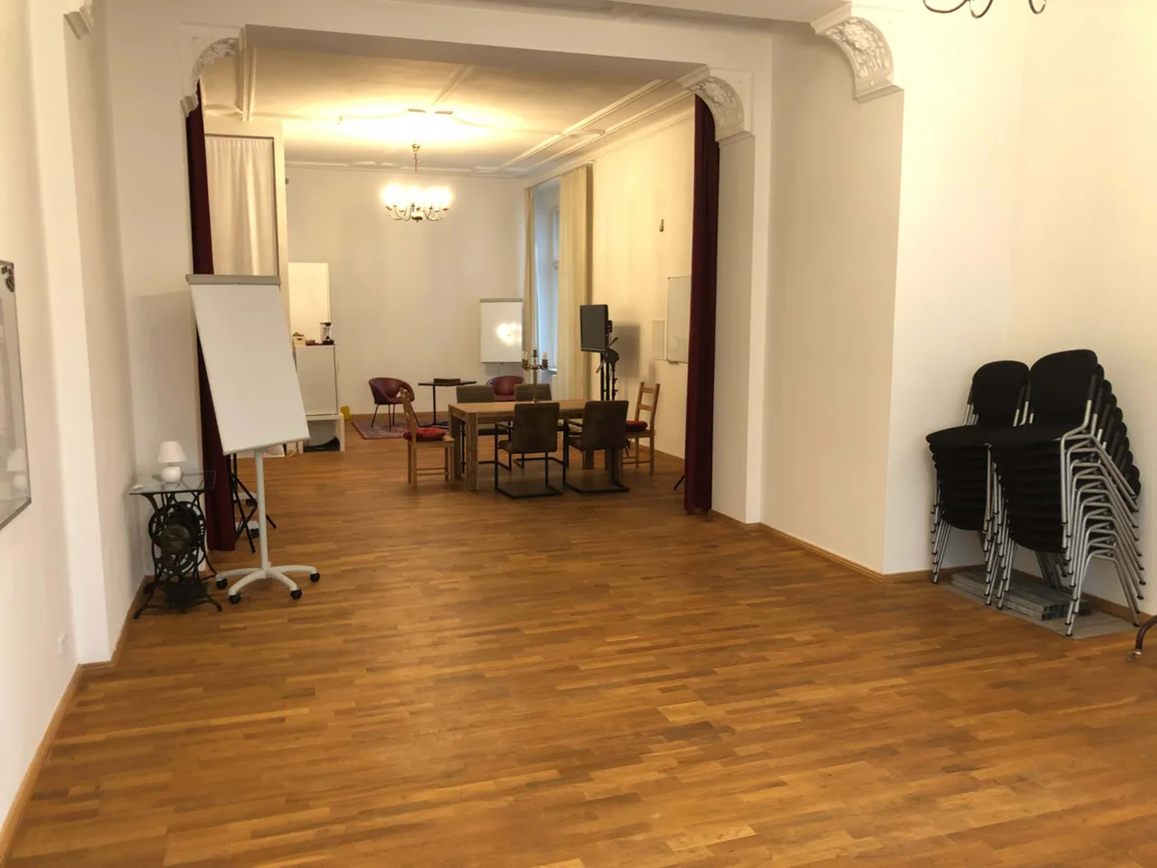 The image shows a spacious, well-lit room with wooden flooring, featuring several chairs and a table in the center, a whiteboard, and stacked chairs in the corner.