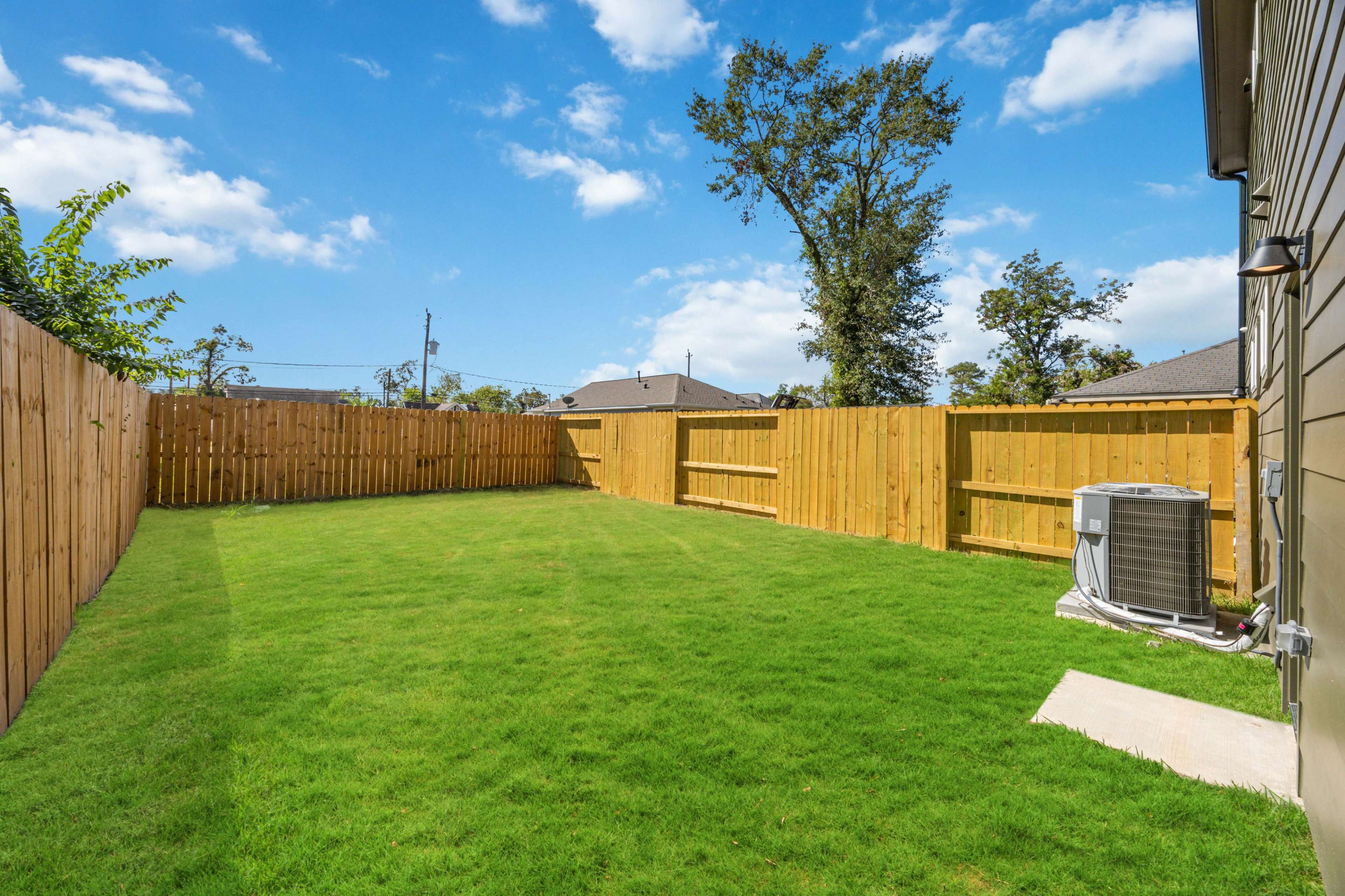 The image shows a spacious backyard enclosed by wooden fences, featuring green grass and an air conditioning unit against a house.