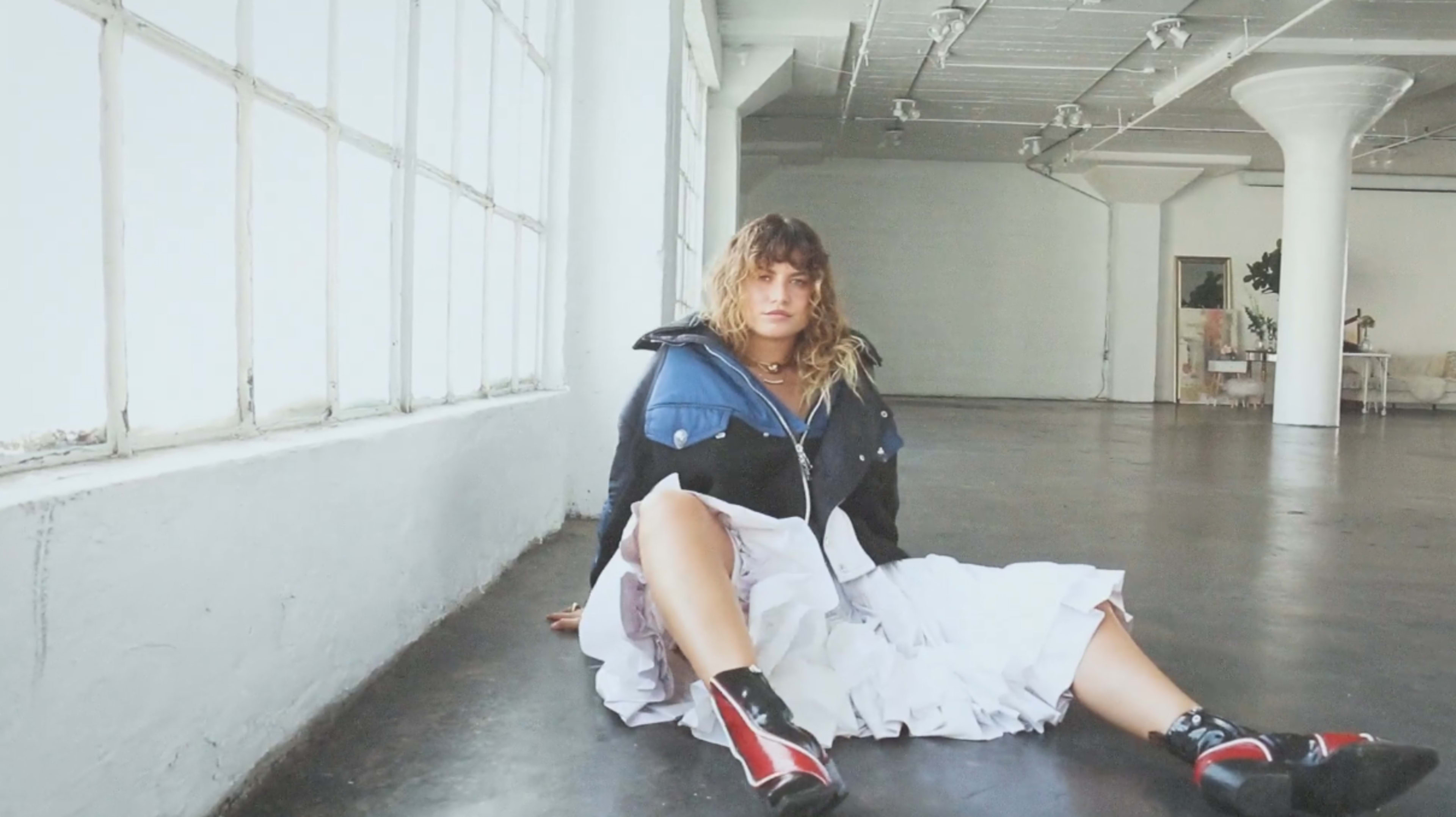 A person is sitting on the floor in an industrial space with large windows, wearing a layered outfit that includes a white dress and a blue jacket.