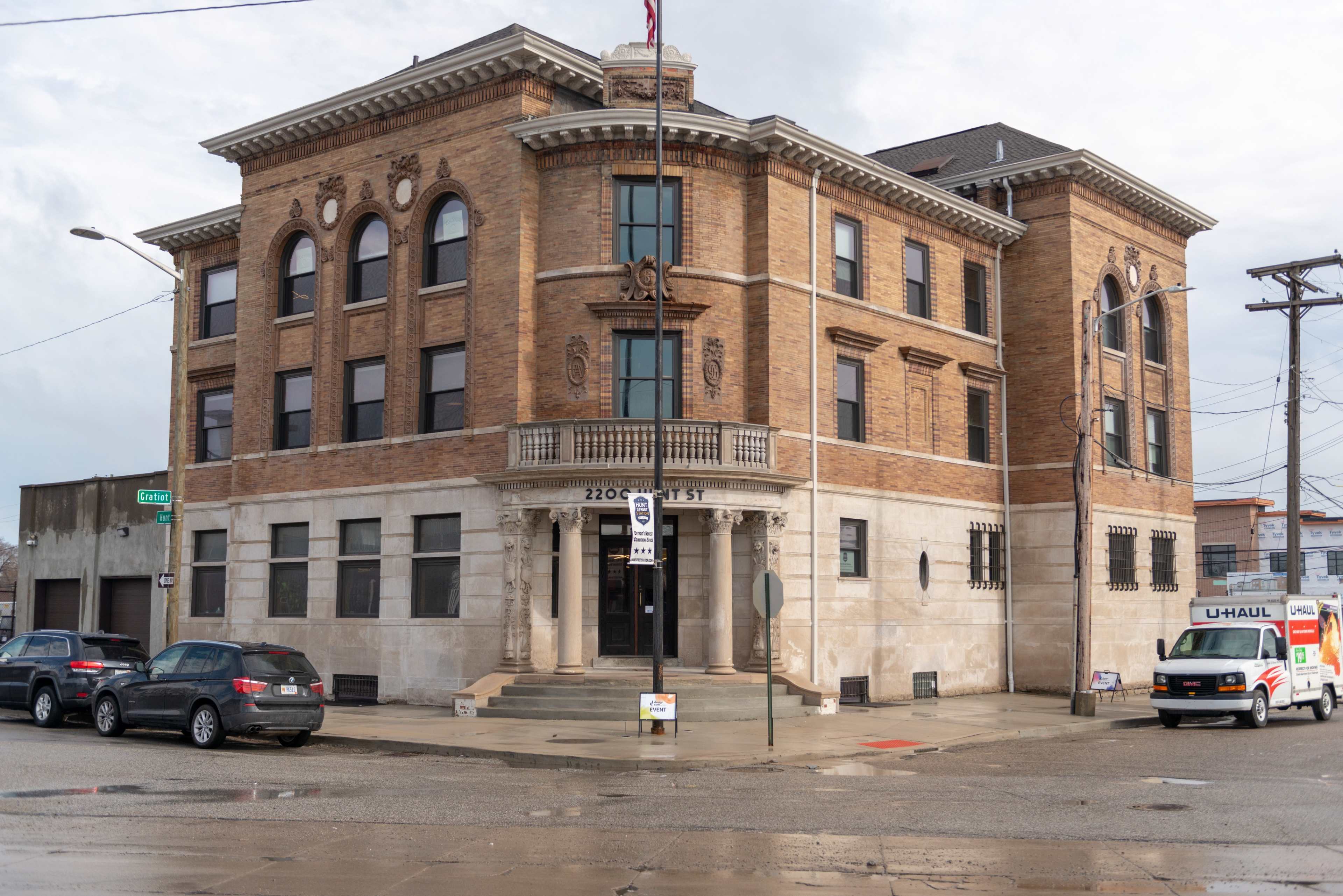A large, ornate brick building with stone details located at the corner of a street, featuring a flagpole and a U-Haul truck parked nearby.