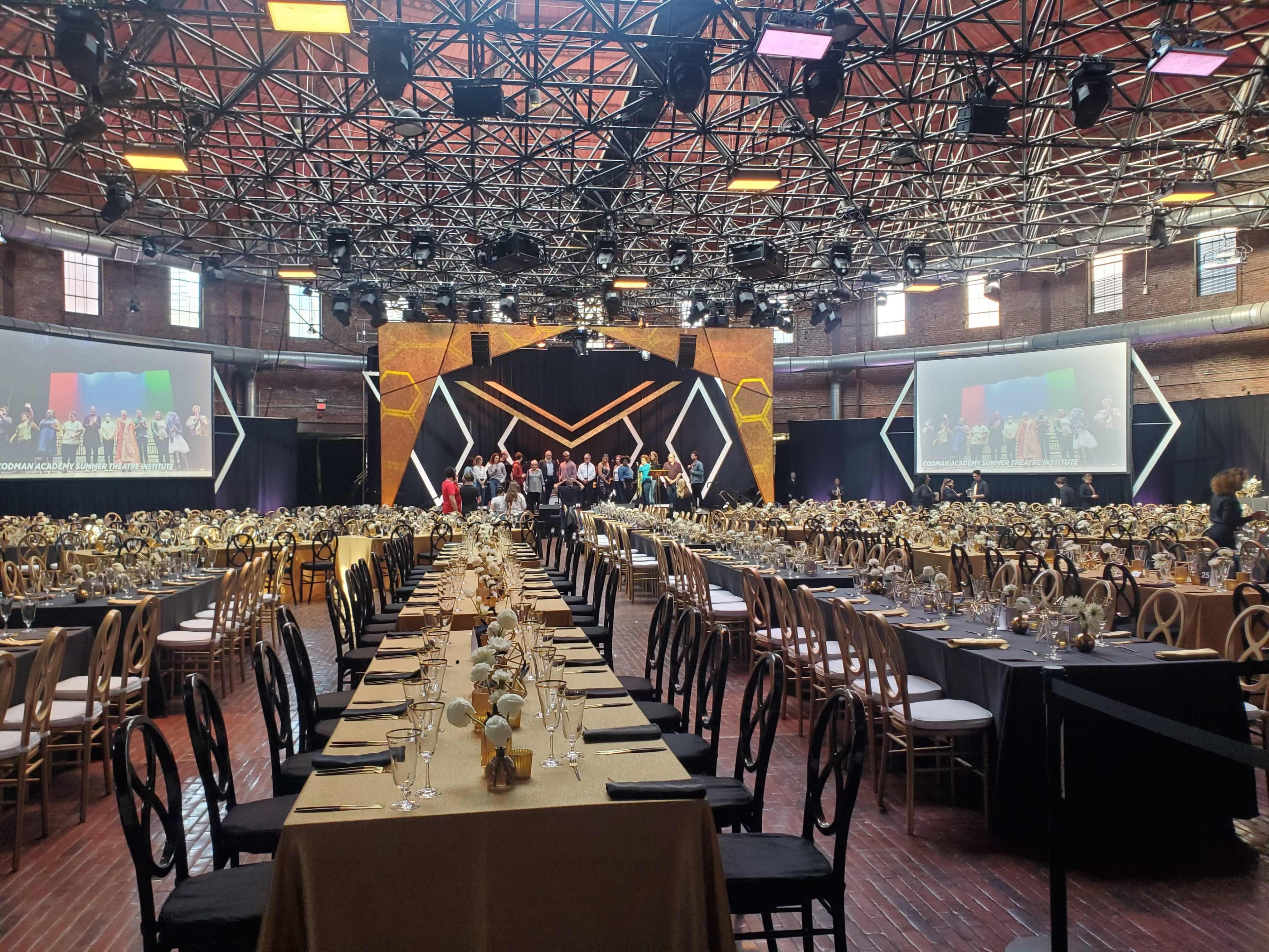 A large banquet hall is set up with elegantly arranged tables, and a stage in the background displays a group of people, with projection screens on either side.