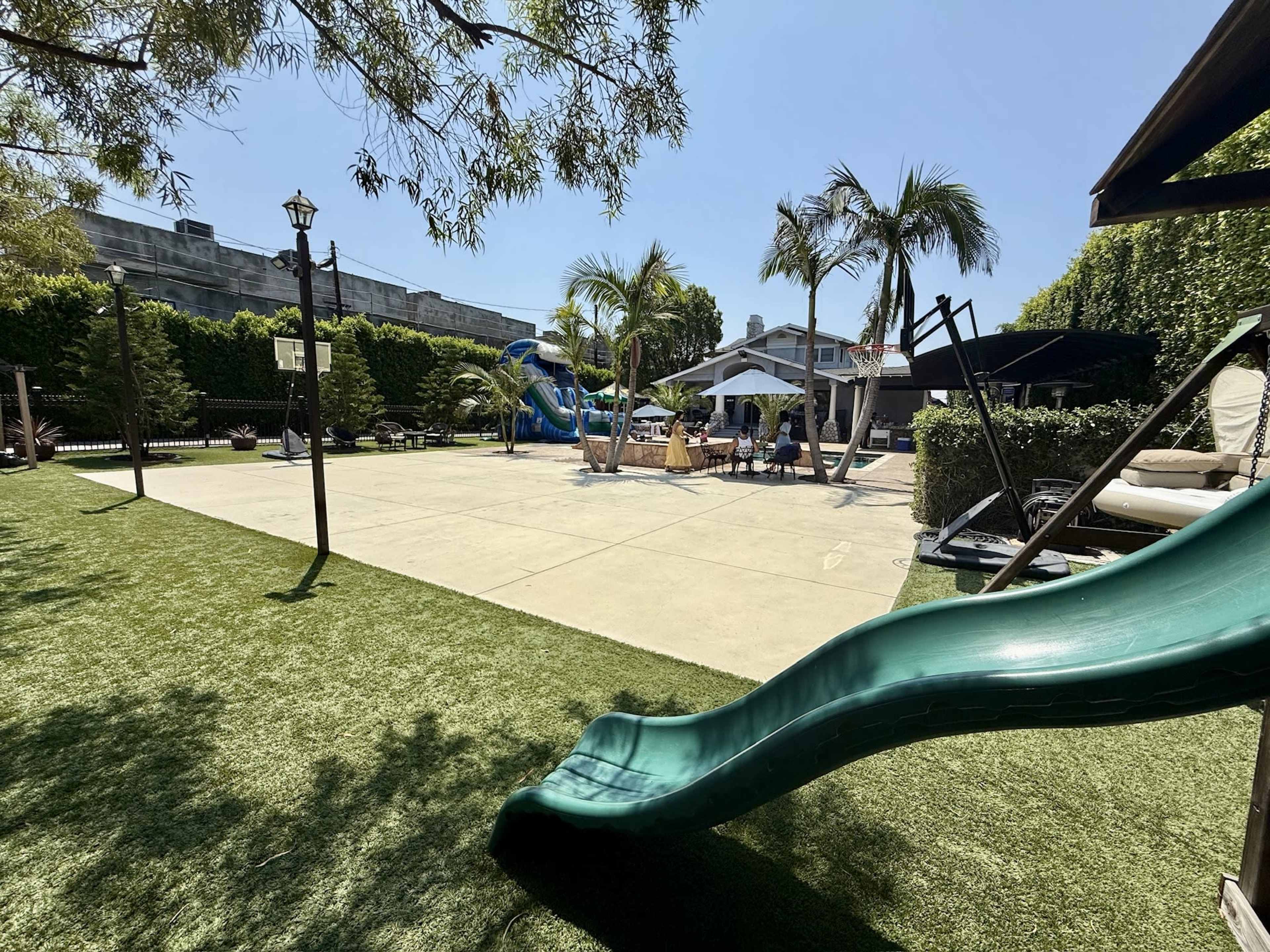 The image shows a backyard with a slide, pool area, lounge chairs, and palm trees under a clear blue sky.