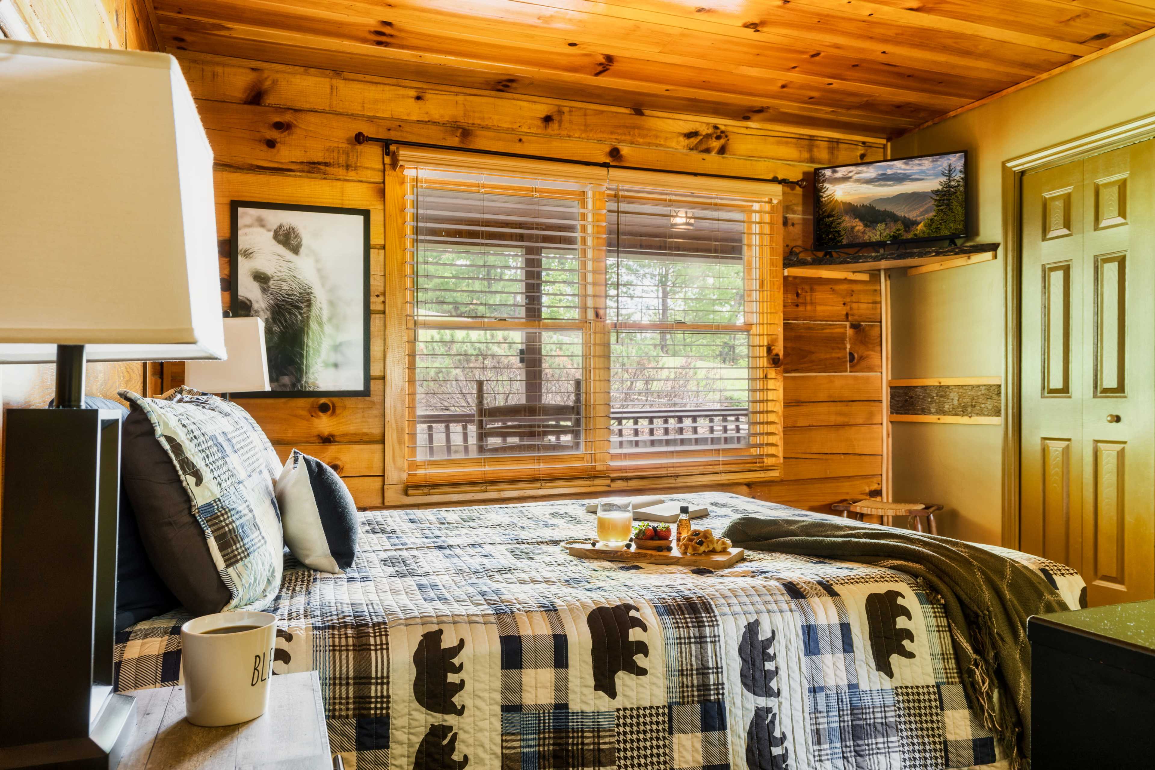 A wooden-paneled bedroom with a plaid bedspread featuring bear designs, a small table set with breakfast items, and a window with blinds letting in natural light.
