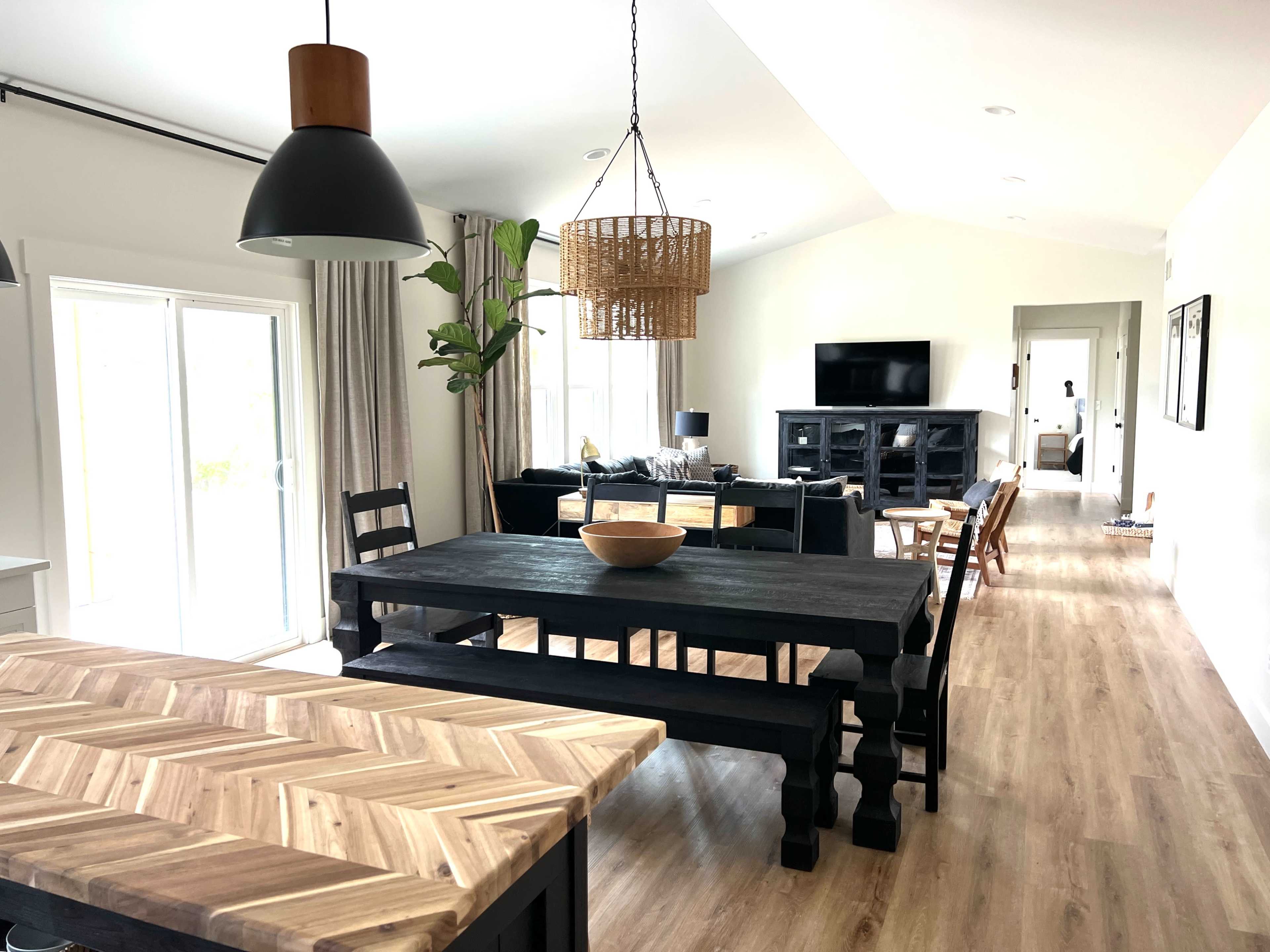 The image shows a modern open-concept dining and living area featuring a large black dining table, a woven pendant light, and a living room with a dark sofa and a television.