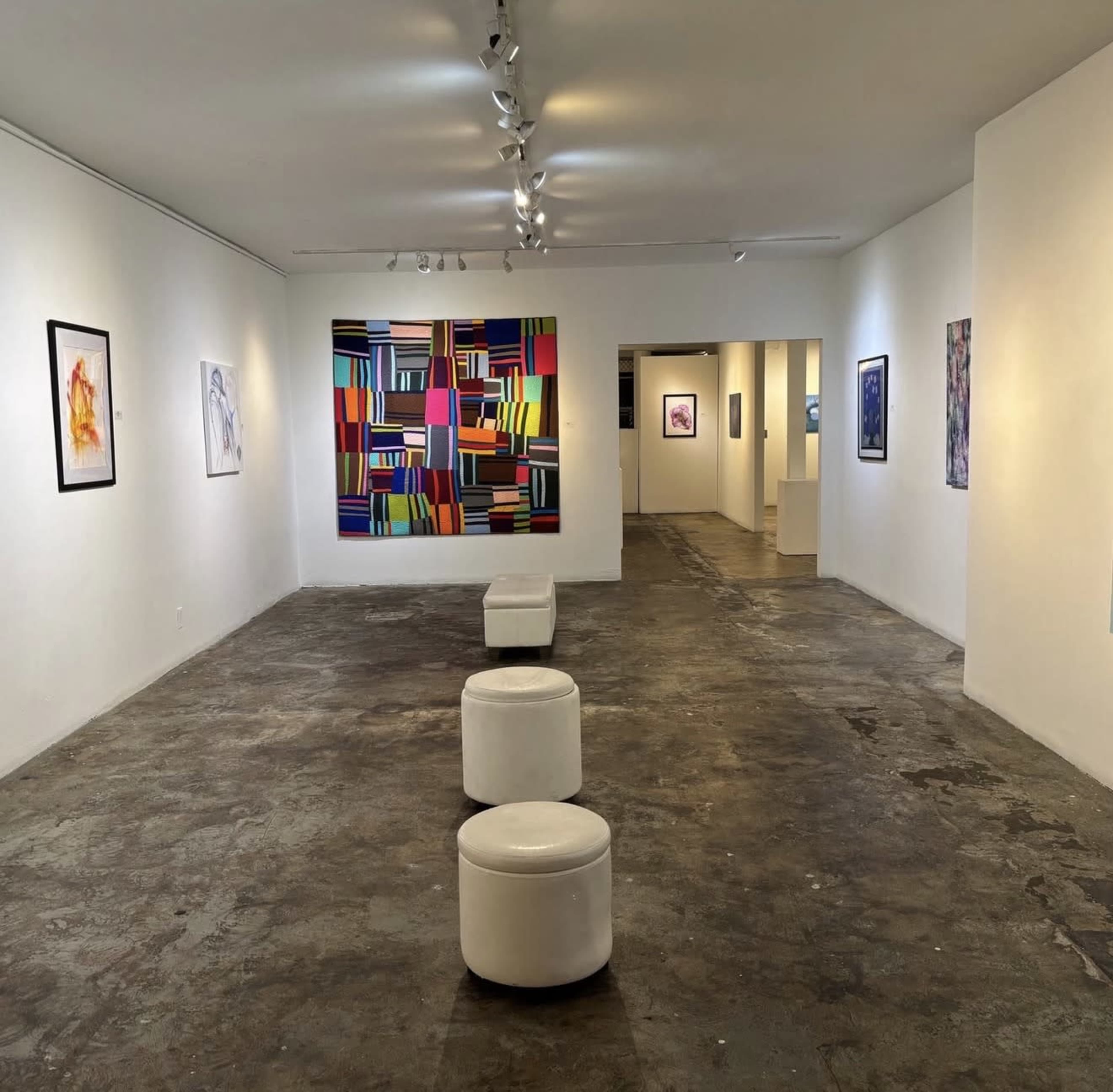 The image shows a modern art gallery with white walls, a colorful abstract painting, and several small white stools arranged along the center of the room.