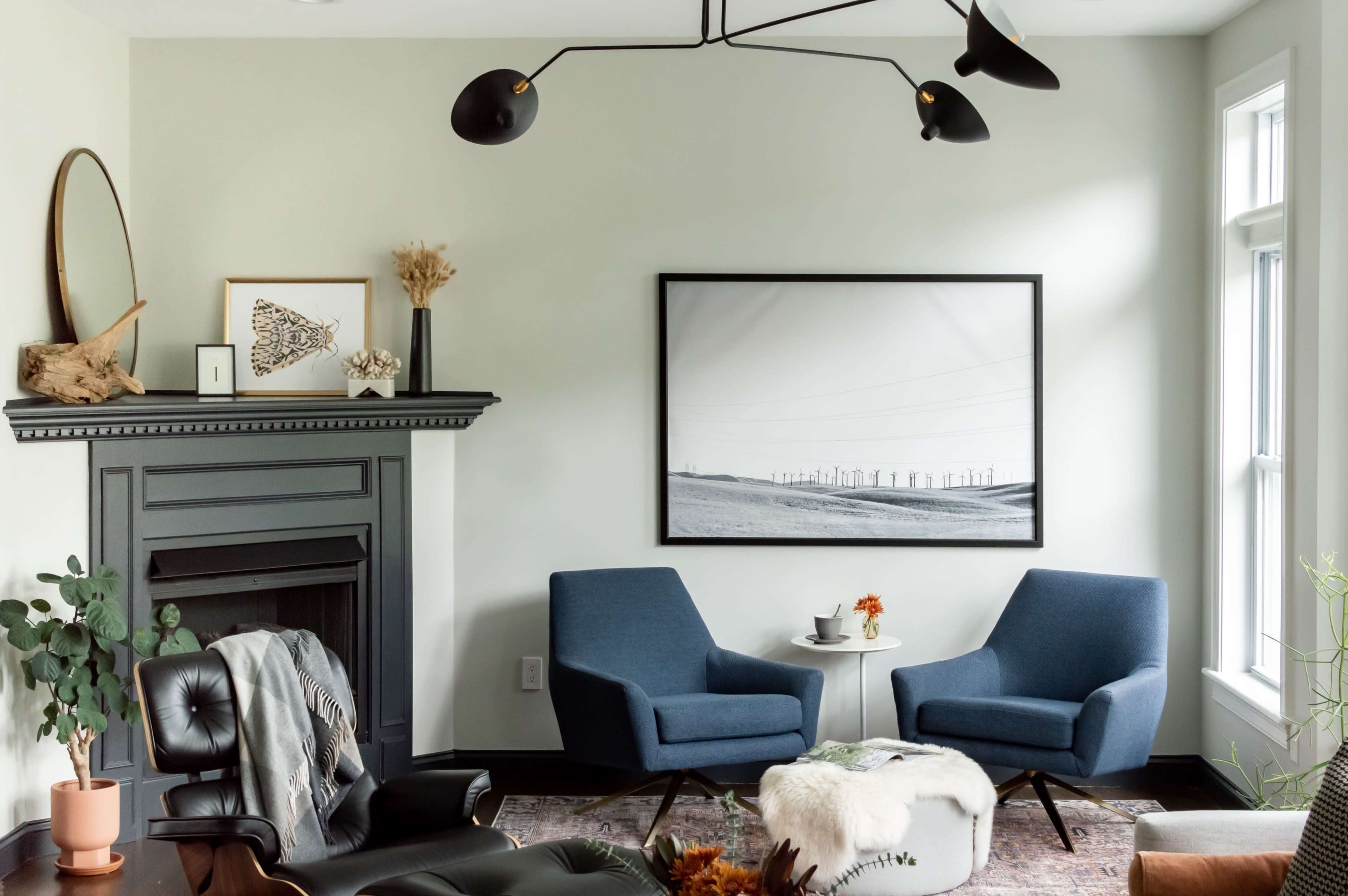 A modern living room features two blue armchairs facing a black-and-white landscape photograph, with a fireplace adorned with decorative items and a plant in the corner.