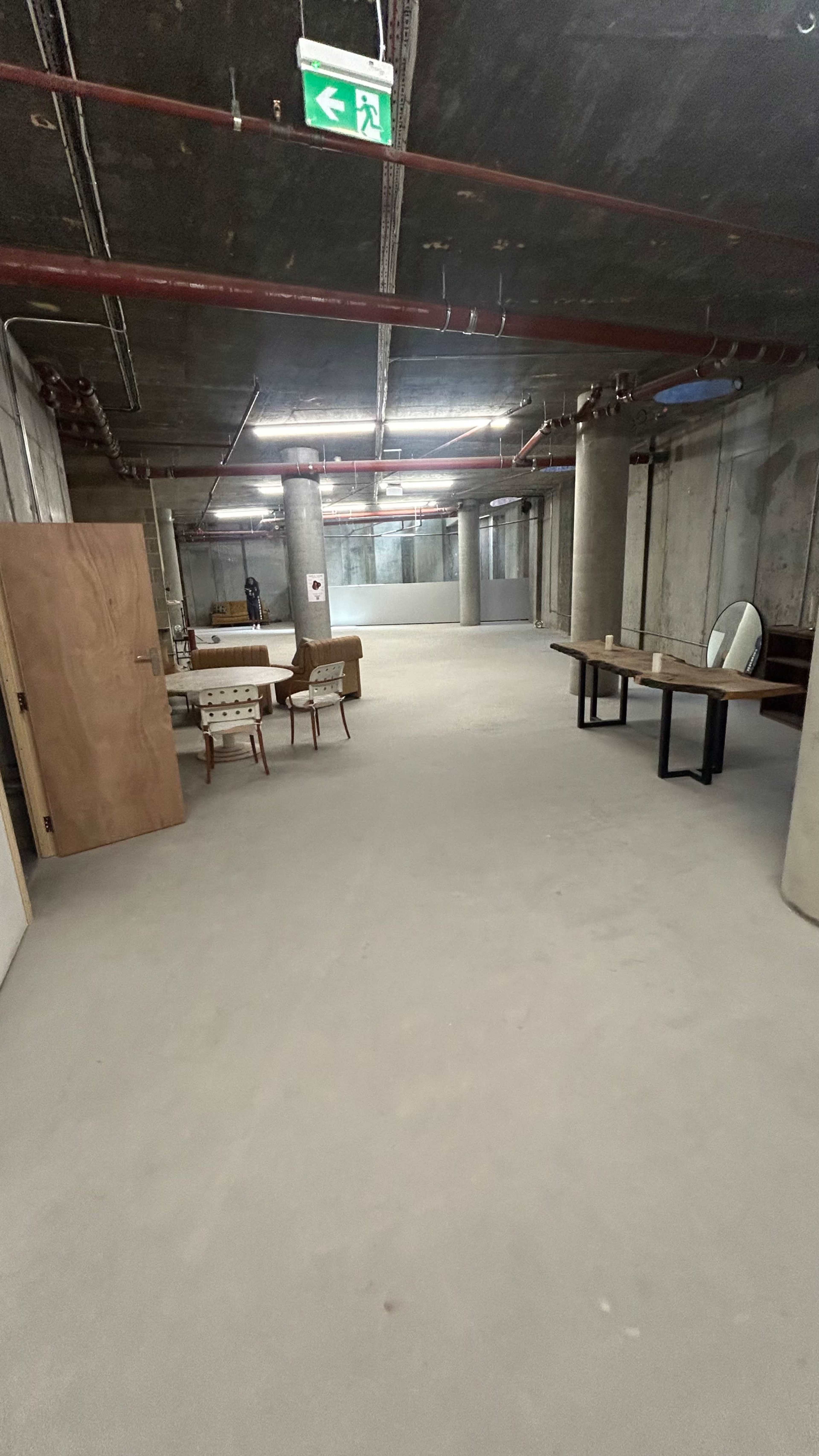 The image shows a sparse, unfinished basement area with concrete walls, exposed pipes, and minimal furniture, including a table and chairs.