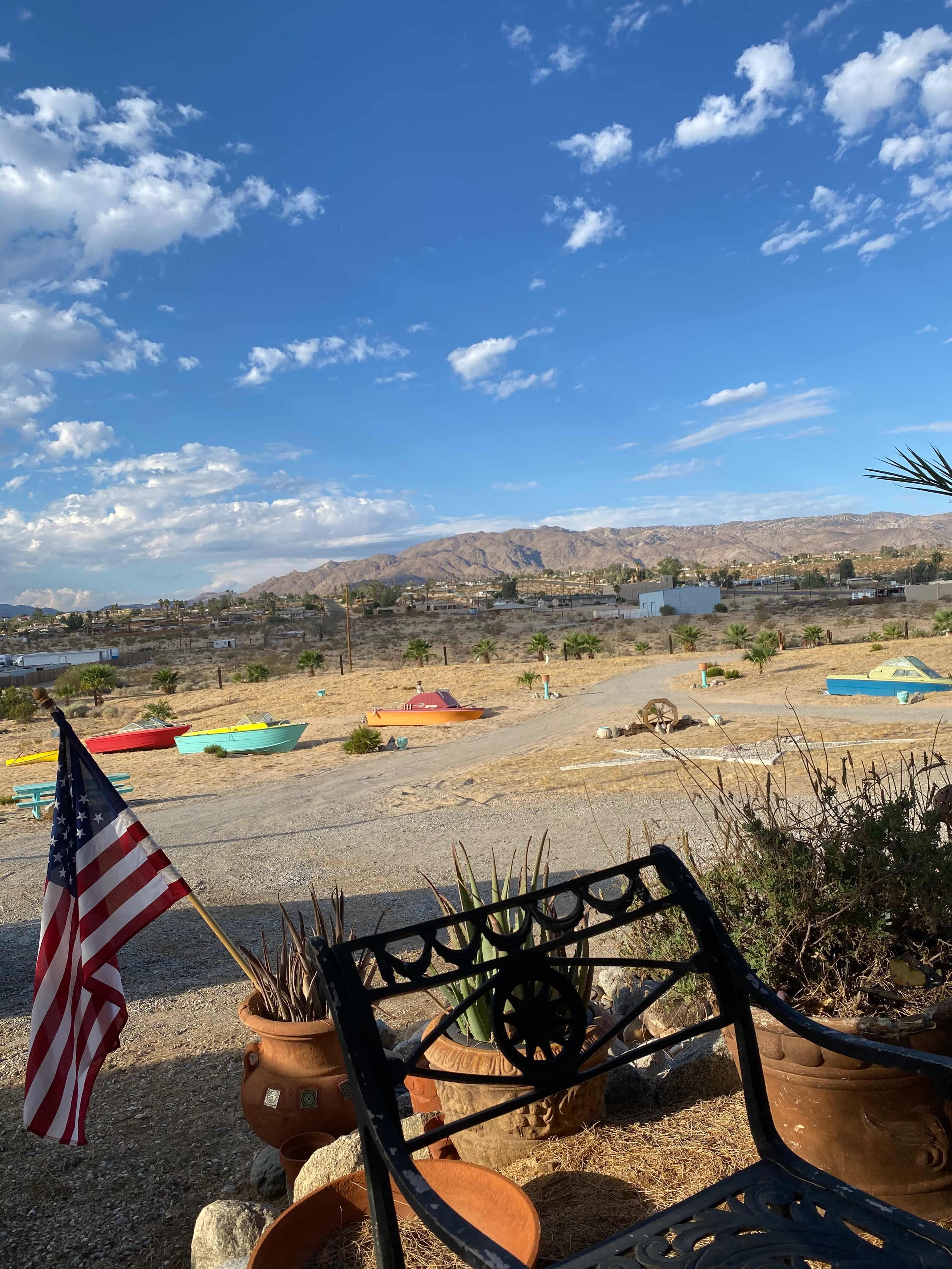 A bench with an American flag is positioned in the foreground, overlooking a landscape with boats and mountains under a partly cloudy sky.