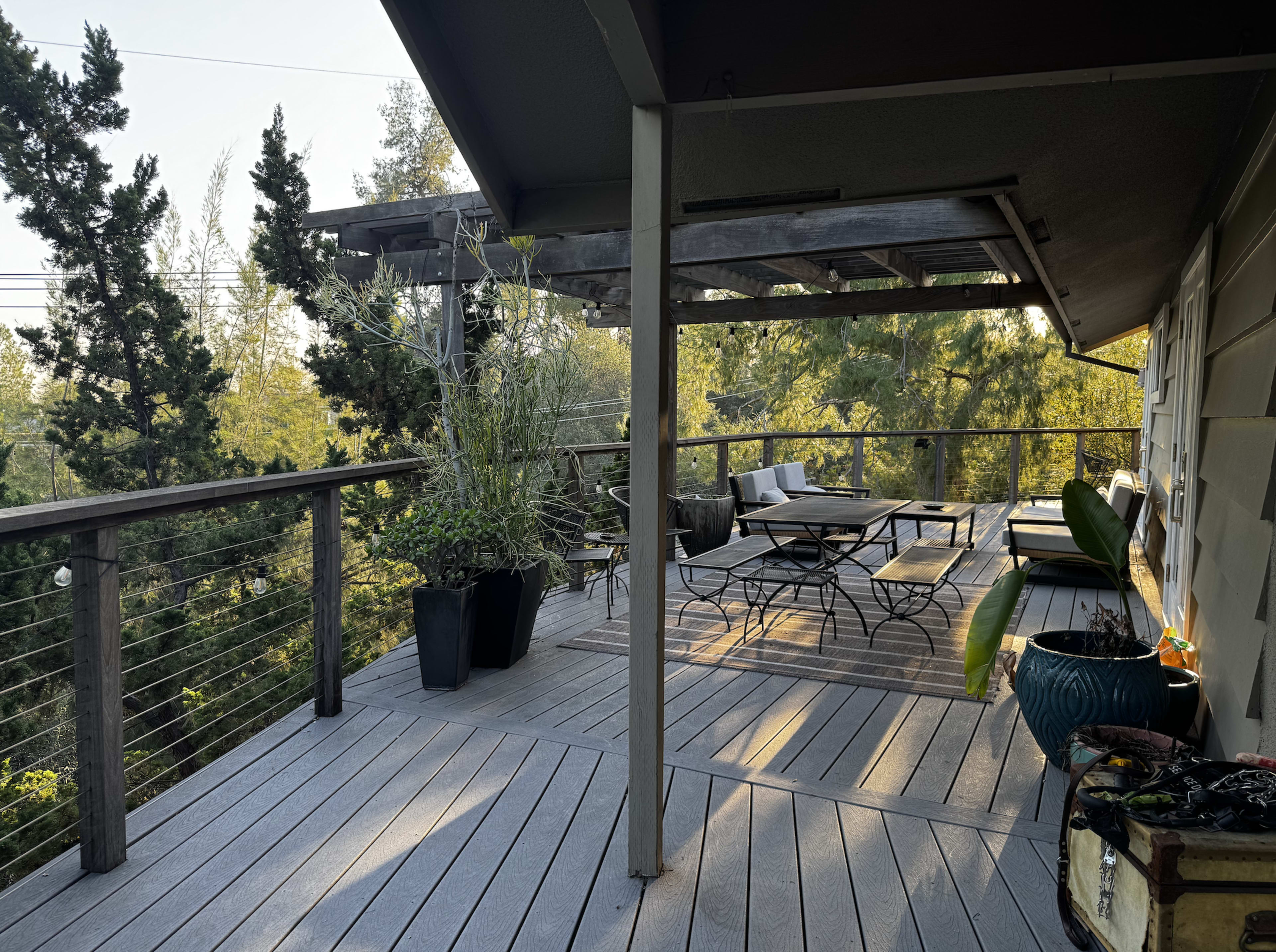 The image shows a wooden deck with a table and chairs, potted plants, and a view of trees in the background.