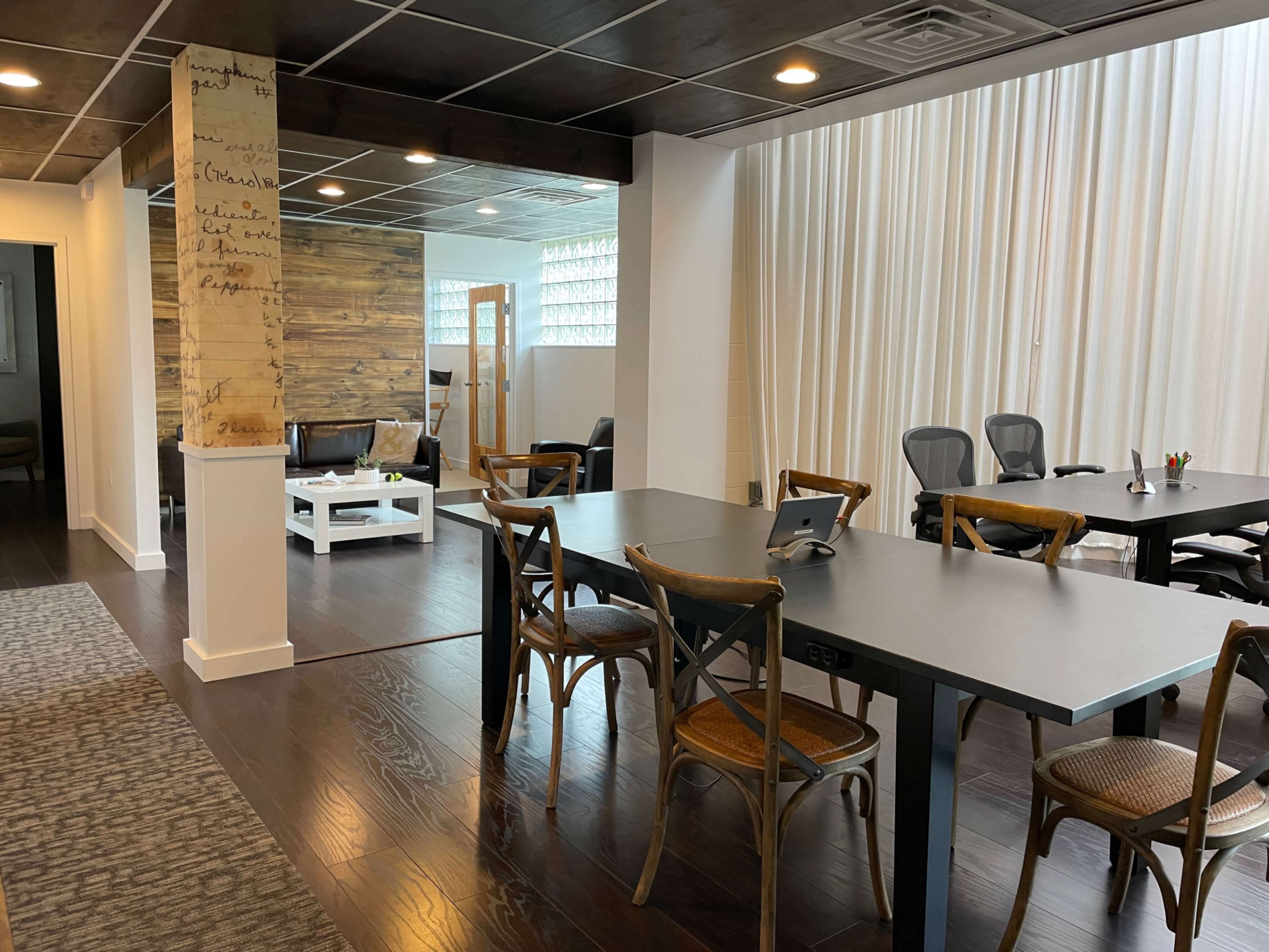 The image shows a modern office space with a long black conference table surrounded by wooden chairs, a seating area with a couch in the background, and large windows featuring white curtains.