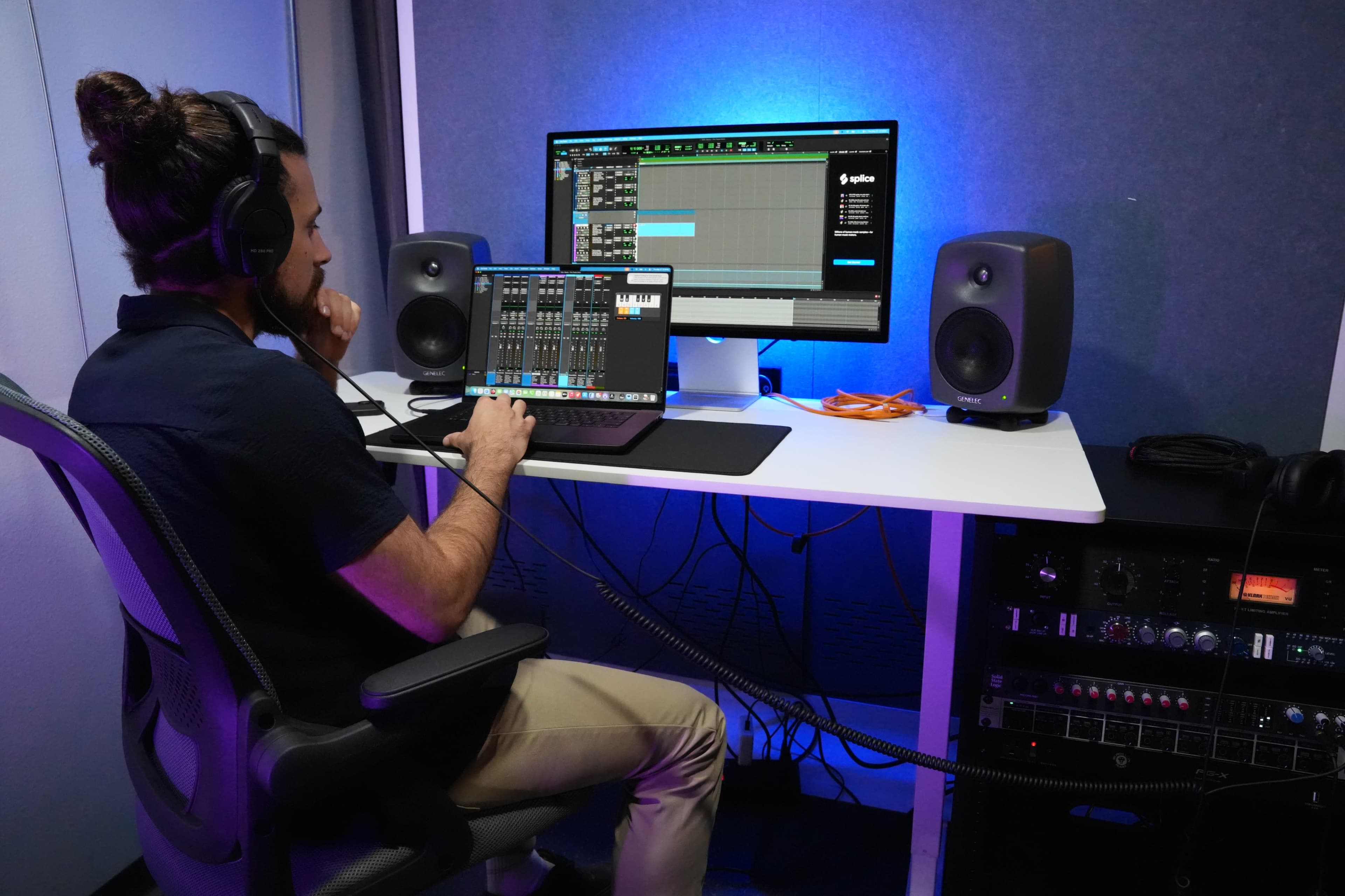 A person sits in a chair at a desk, using a laptop to create music while monitoring audio on a computer screen and speakers.