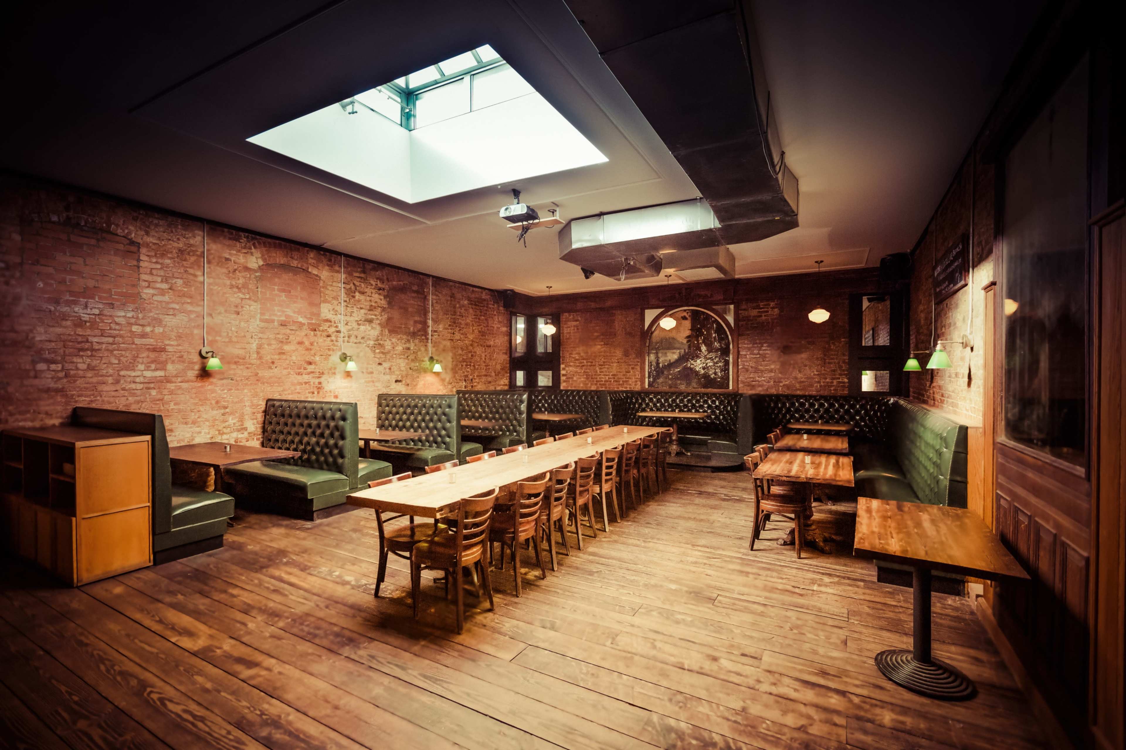 A spacious dining area with wooden floors, brick walls, and a skylight, featuring long tables and green upholstered booths along the walls.