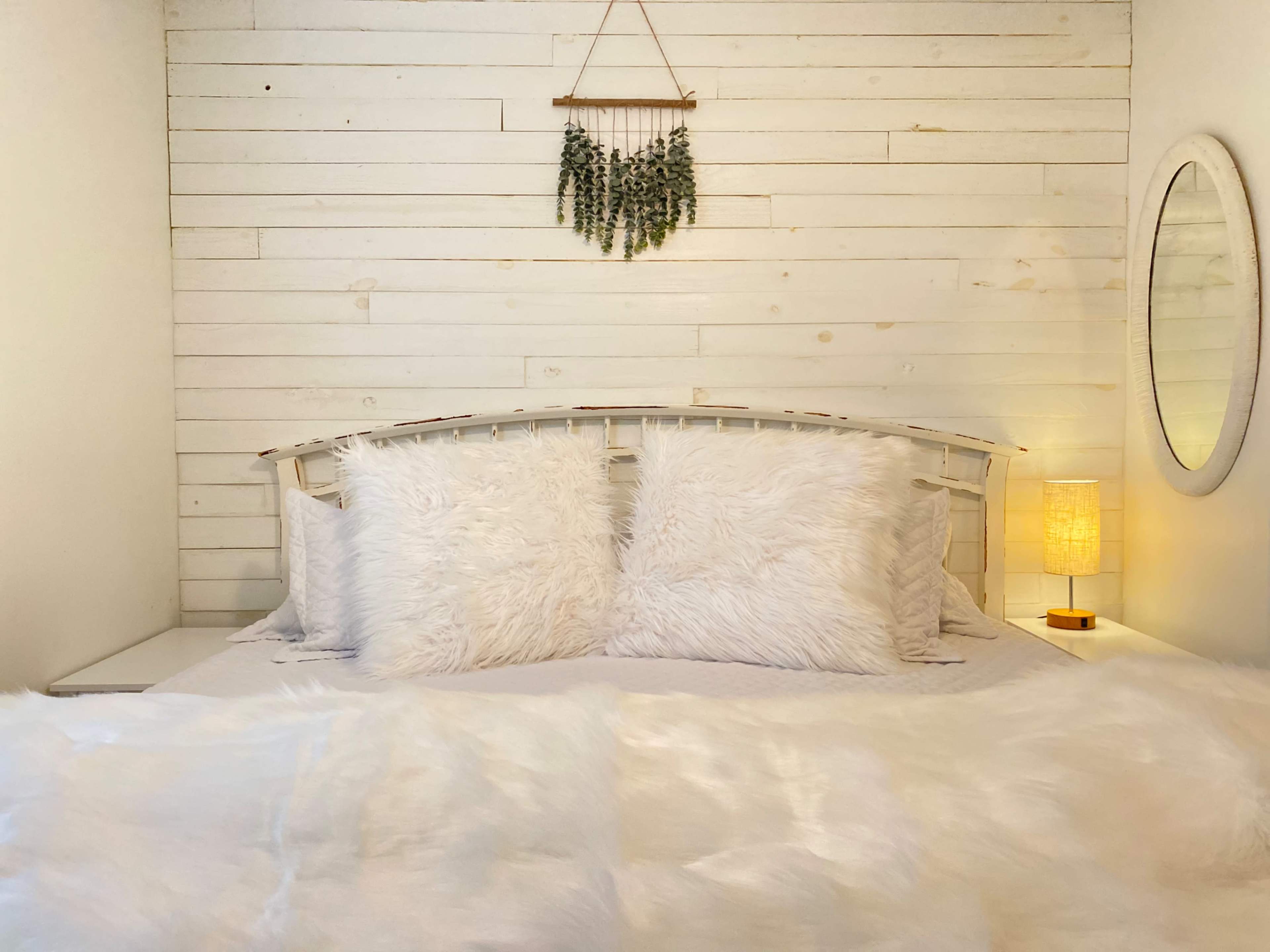 A cozy bedroom with a white wooden headboard, fluffy pillows, and a lamp beside a round mirror on a light-colored wall.