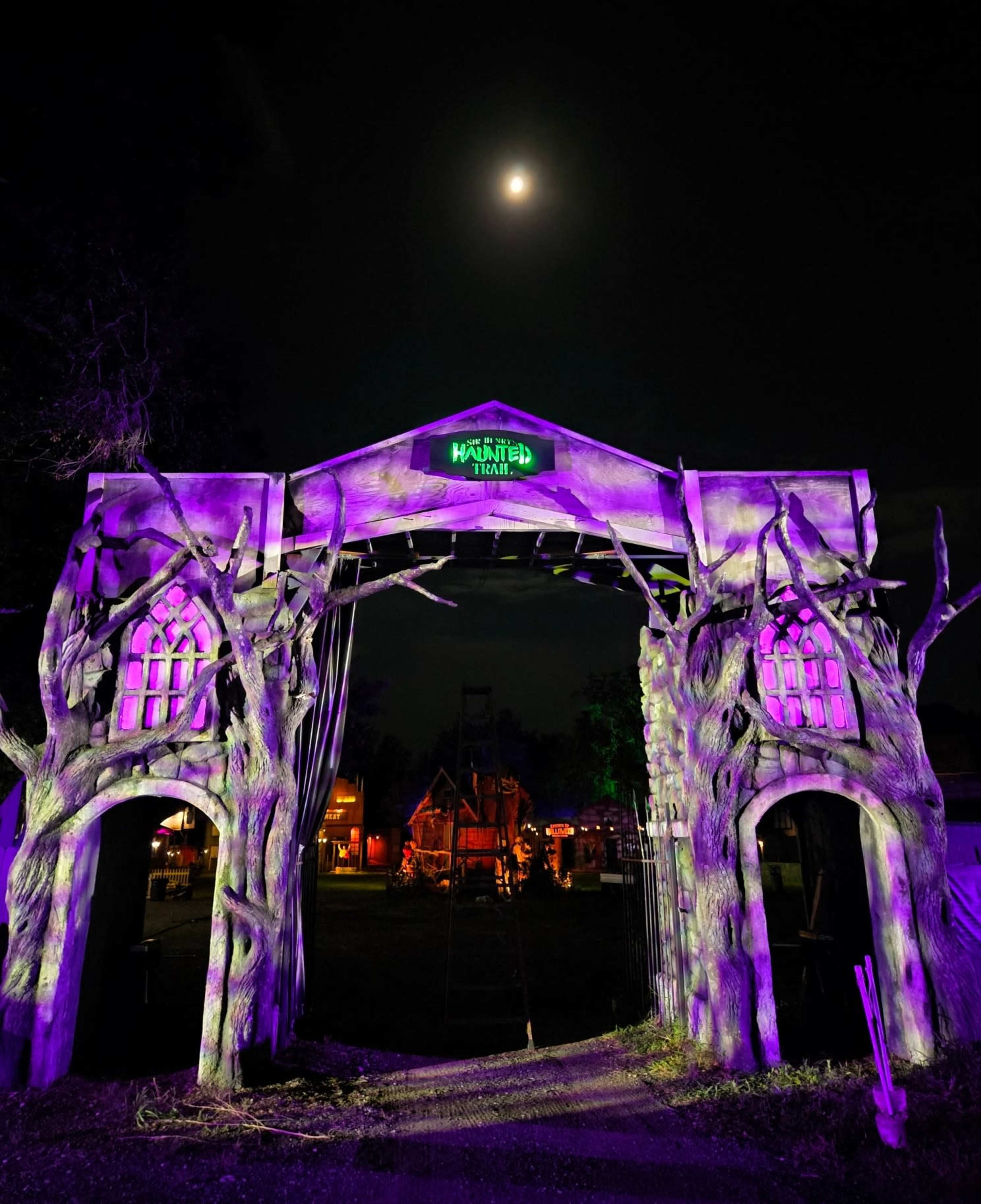 A haunted attraction entrance is illuminated by purple lights under a full moon, featuring twisted trees and gothic arches.