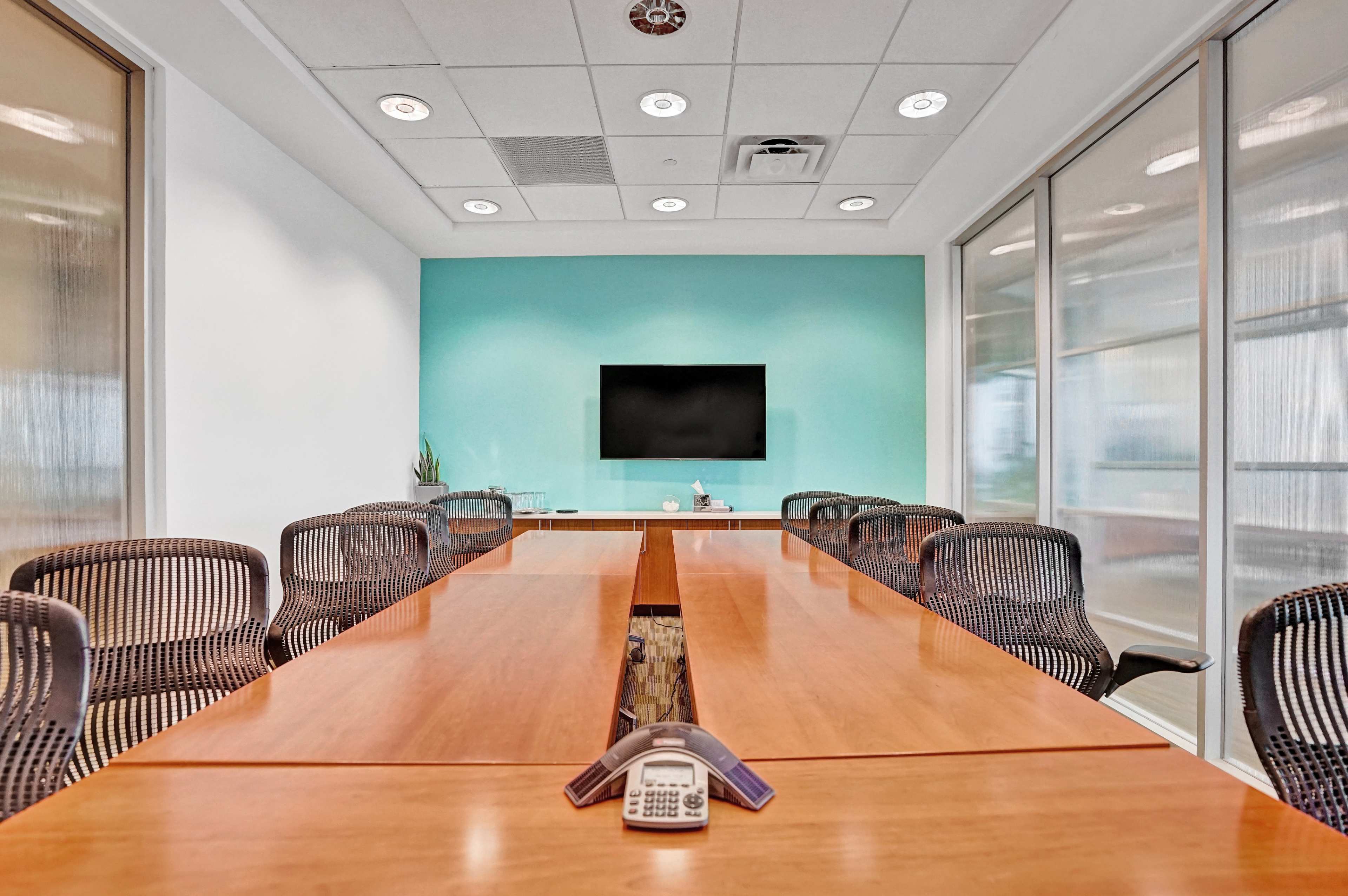 A conference room features a long wooden table surrounded by mesh chairs, with a television mounted on a light blue wall.