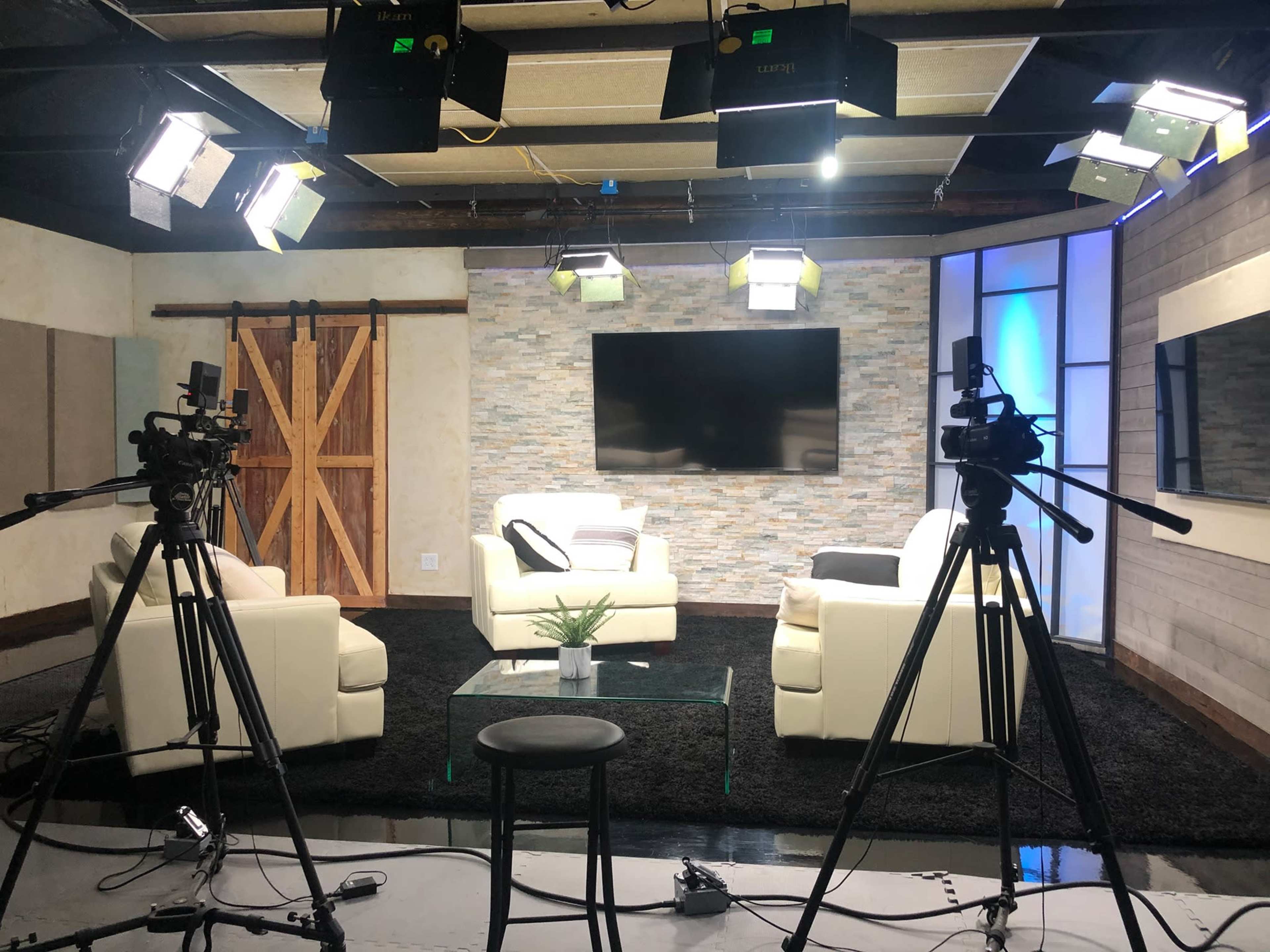 A television studio setup features two white sofas, a glass coffee table, and multiple cameras positioned around a well-lit set with a stone accent wall and a large screen.