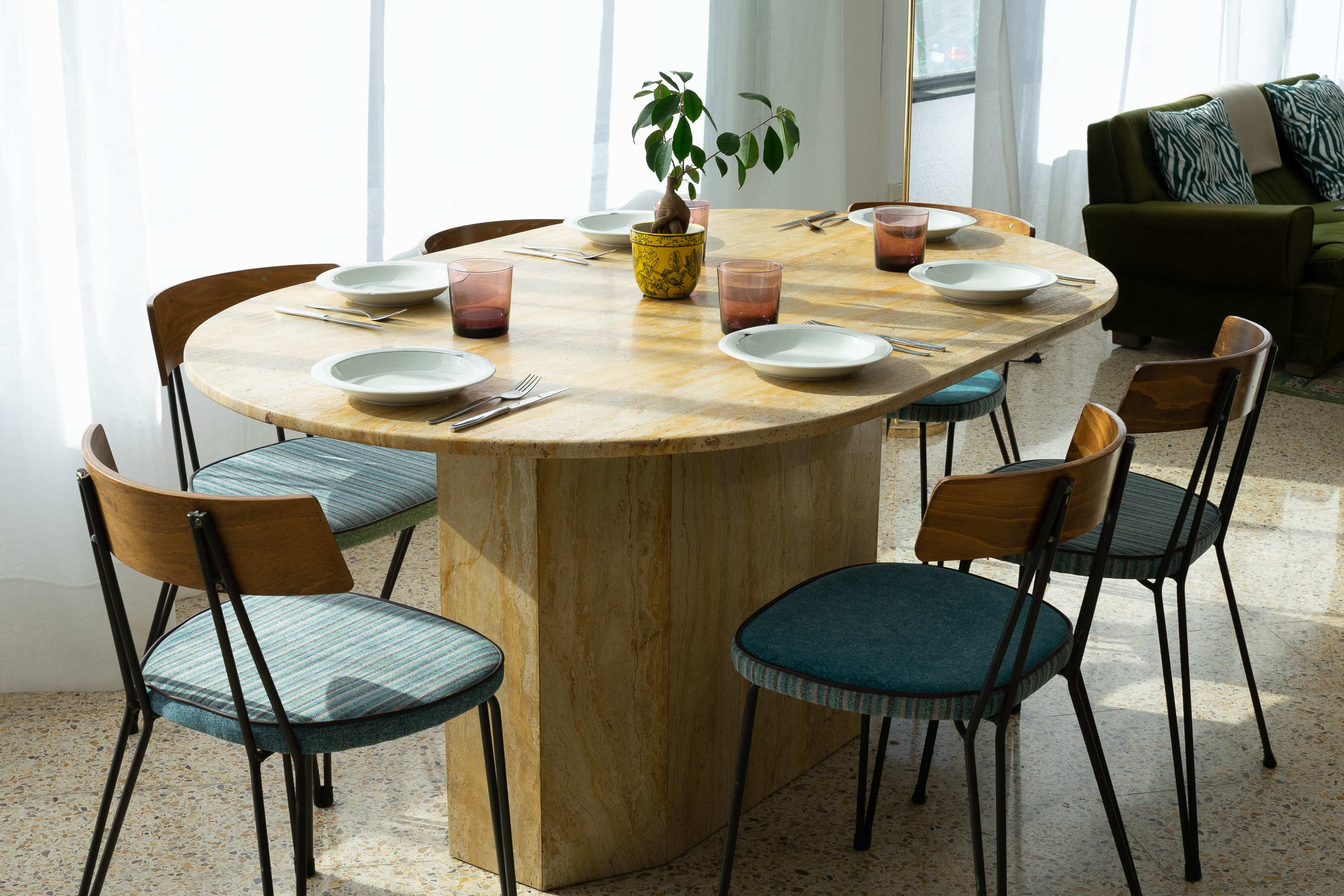A round table with cream-colored stone and wooden accents is set with six plates and glasses, surrounded by chairs, and features a small potted plant in the center.