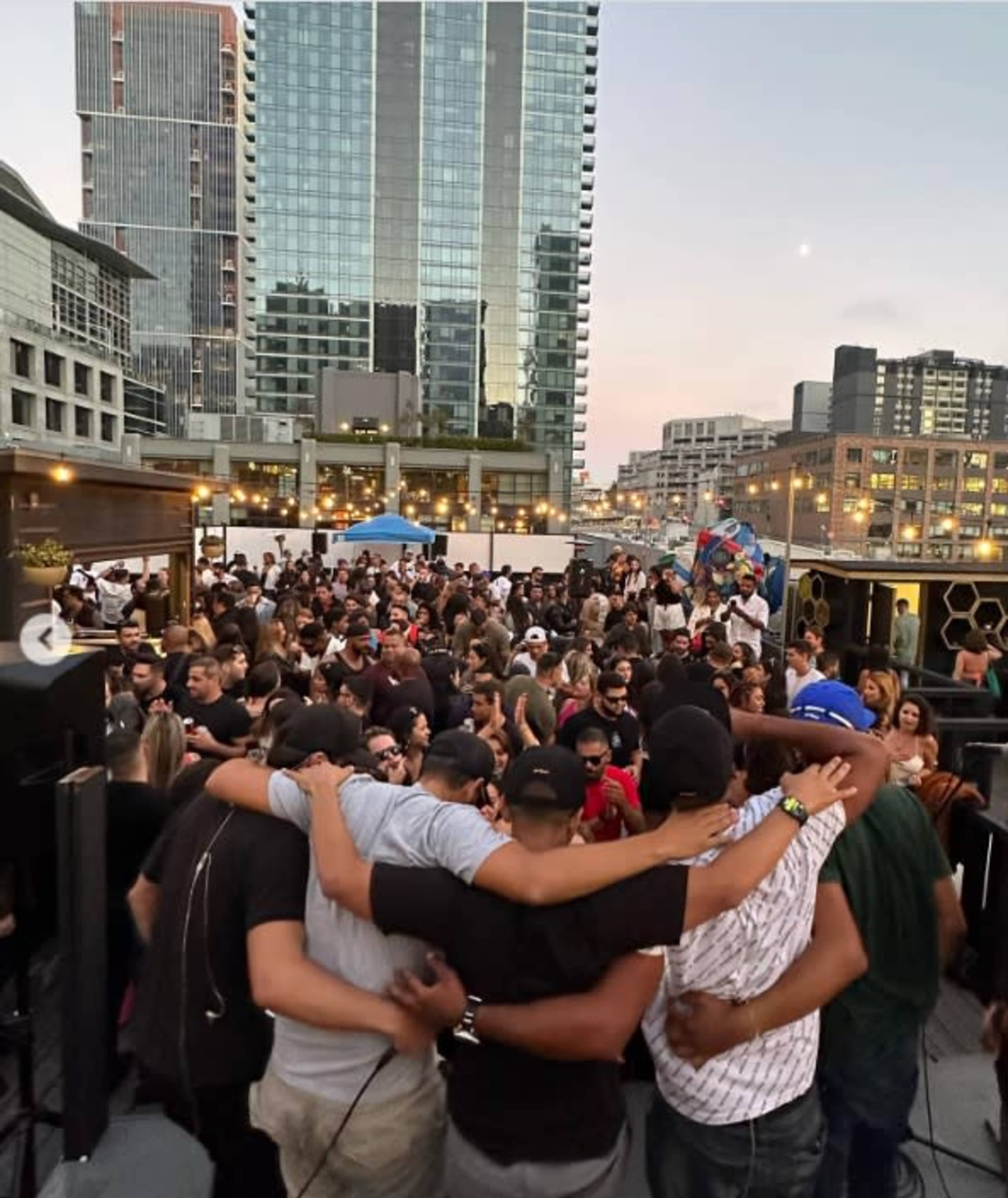 A large group of people gathers on a rooftop in an urban setting, with skyscrapers visible in the background during sunset.