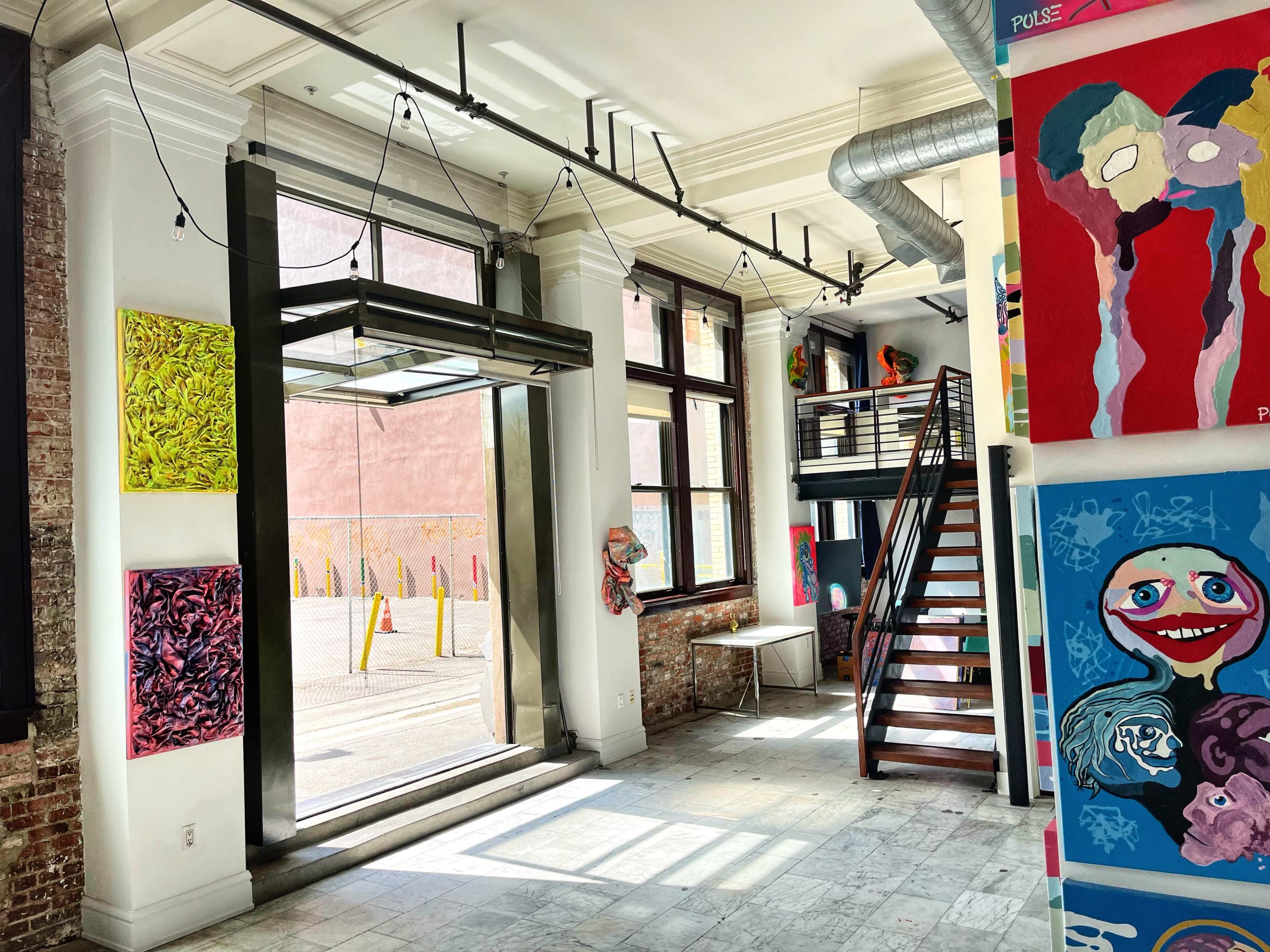 The image shows an art gallery interior featuring a staircase, large windows, and colorful artworks displayed on the walls.