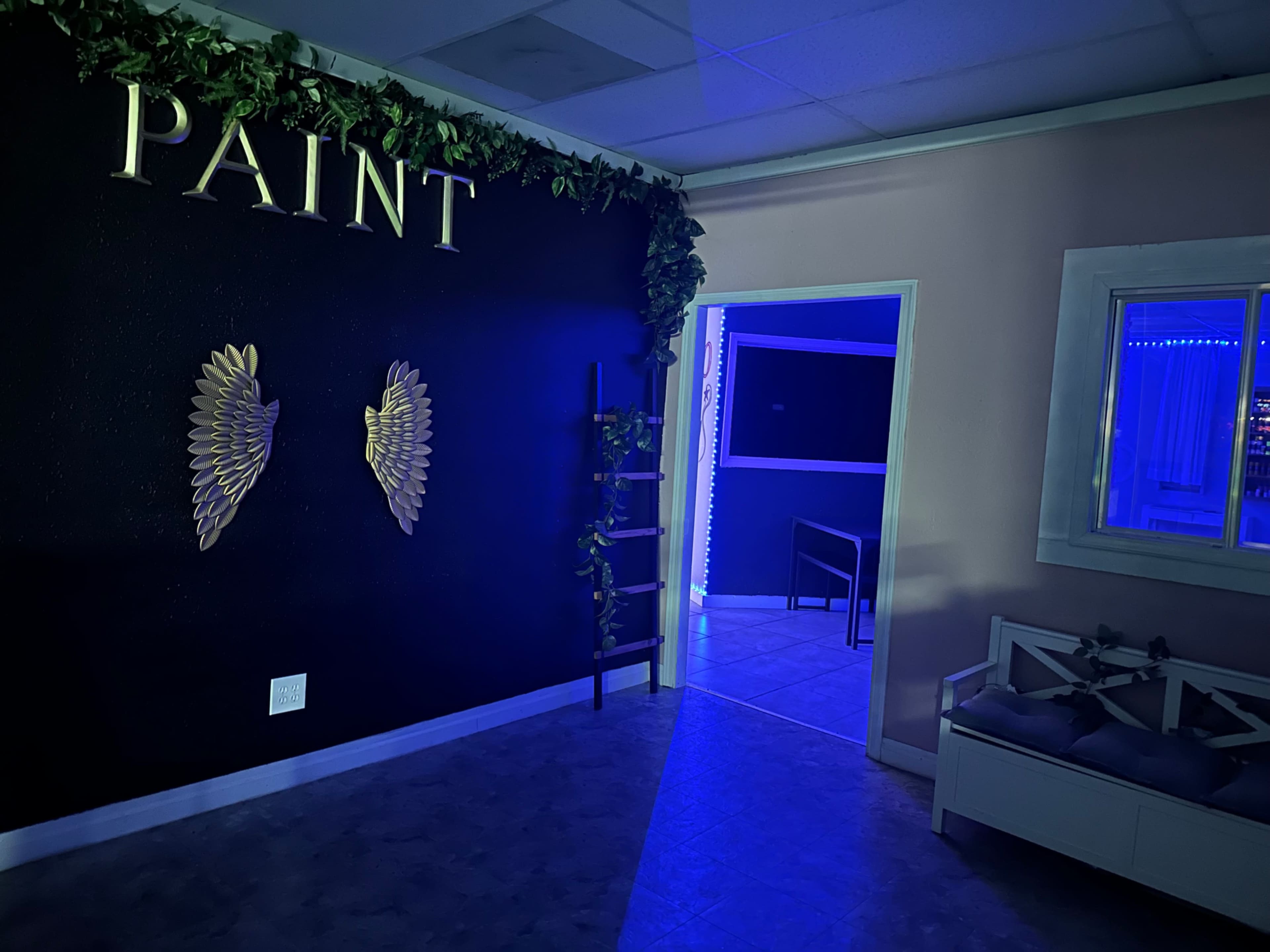 An interior room features a black wall with the word "PAINT" above two decorative wing-shaped wall pieces, and blue lighting illuminating a doorway leading to another space.