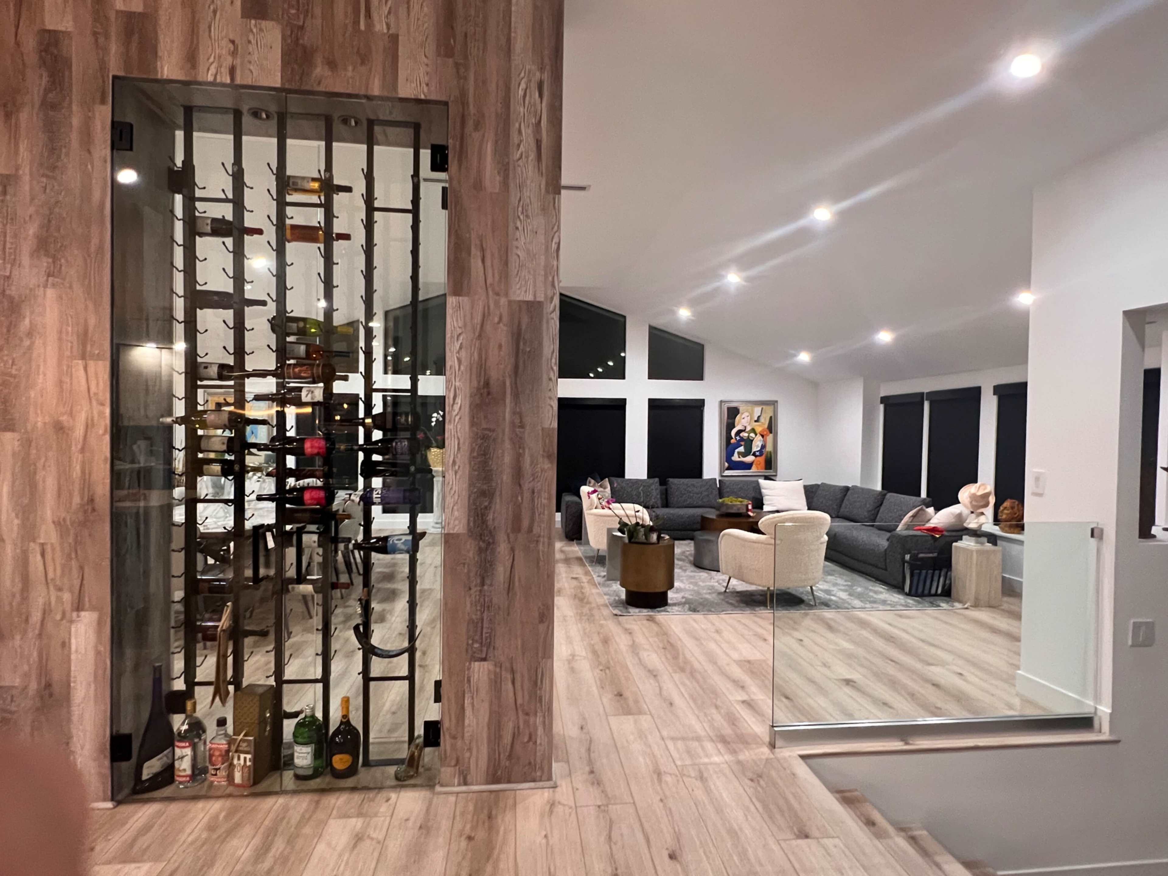 The image shows a modern interior space featuring a wine rack with glass doors, a cozy living room with a sectional sofa, and large windows.