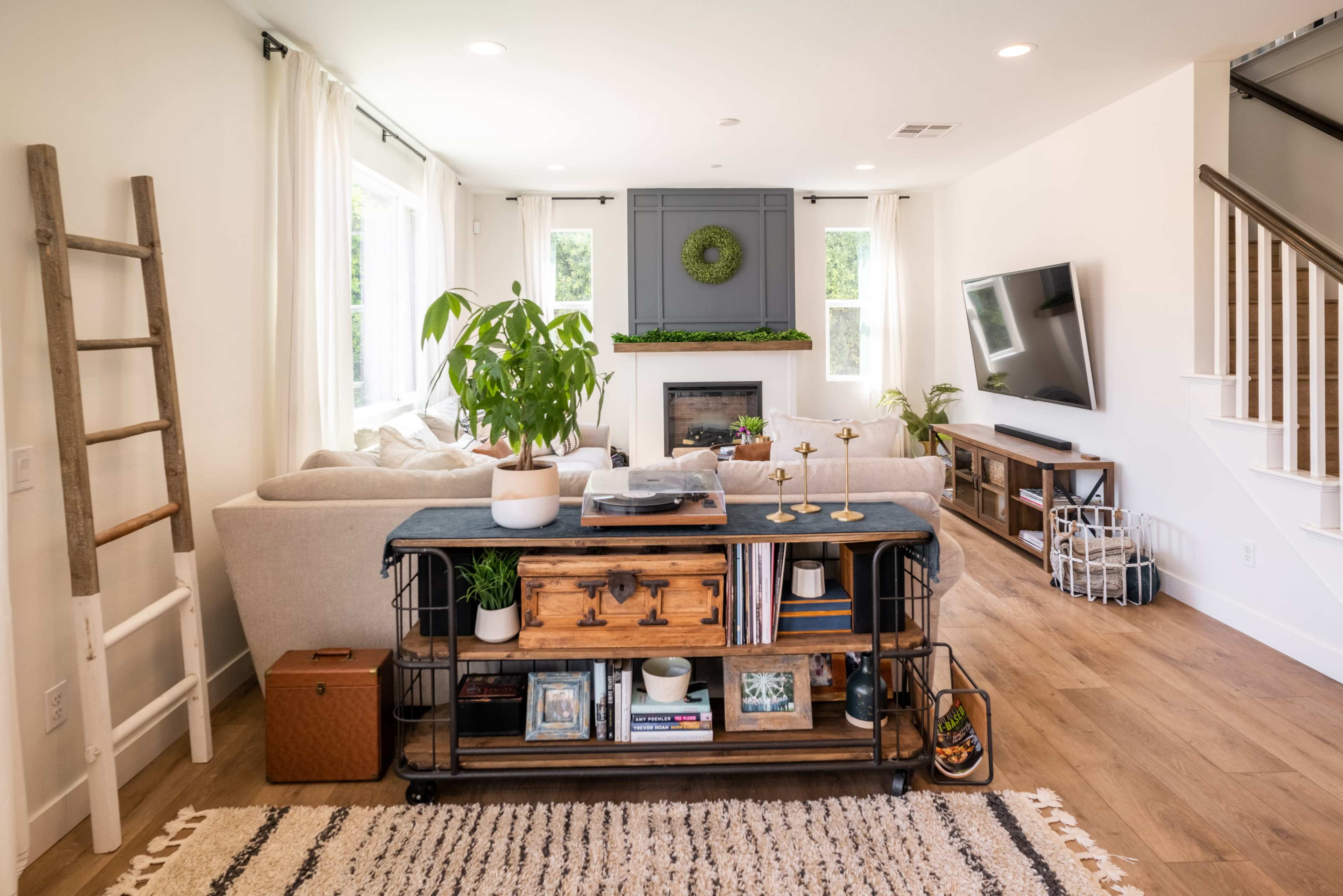 A living room features a light-colored sofa, a wooden coffee table with books and decorative items, and a large TV mounted on the wall, with windows allowing natural light.