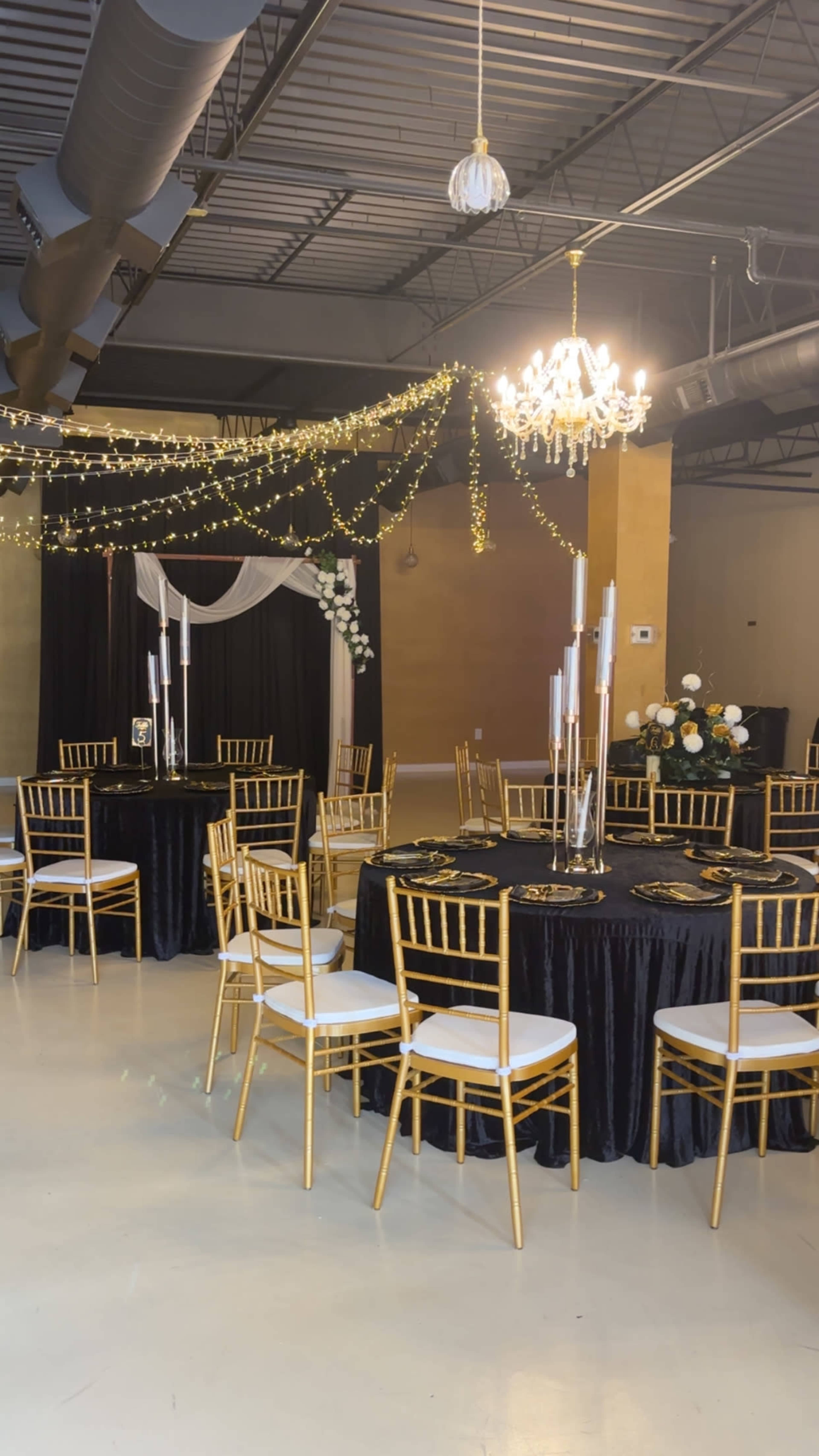 The image shows an elegantly decorated event space with round black tables, gold chairs, and a chandelier adorned with string lights and floral arrangements.
