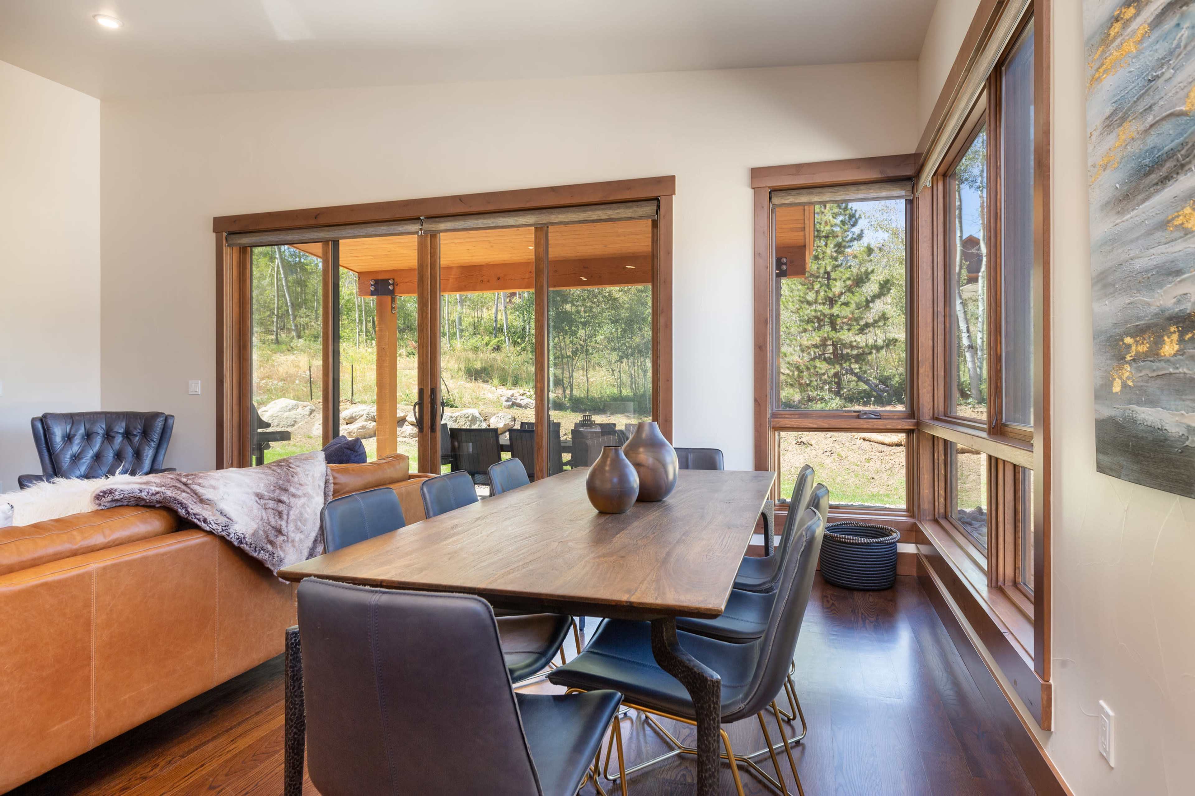 A modern dining area features a wooden table surrounded by black chairs, with large windows showcasing a view of trees and outdoor seating.