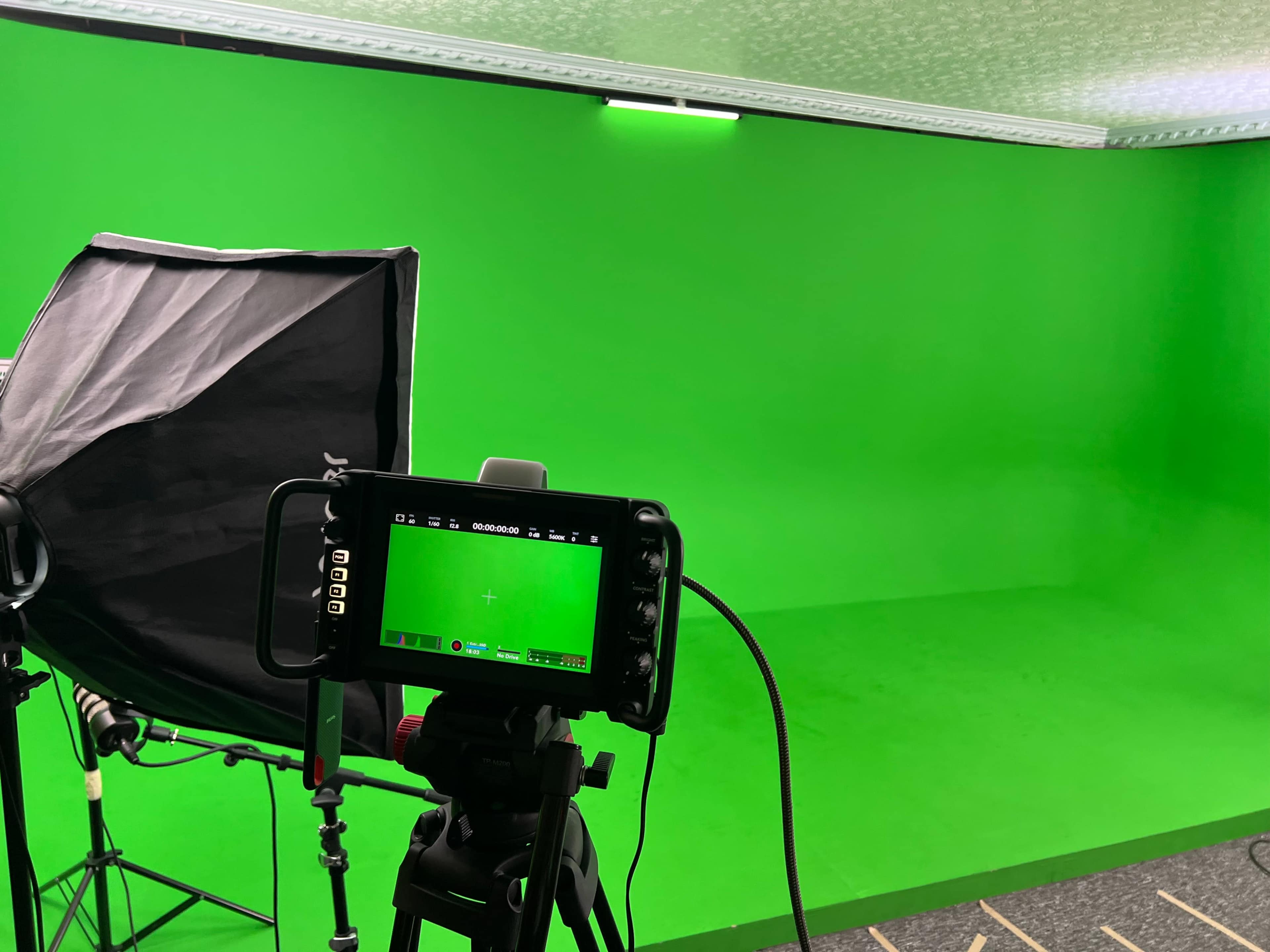 The image shows a green screen setup with a camera on a tripod and a softbox light in a studio setting.